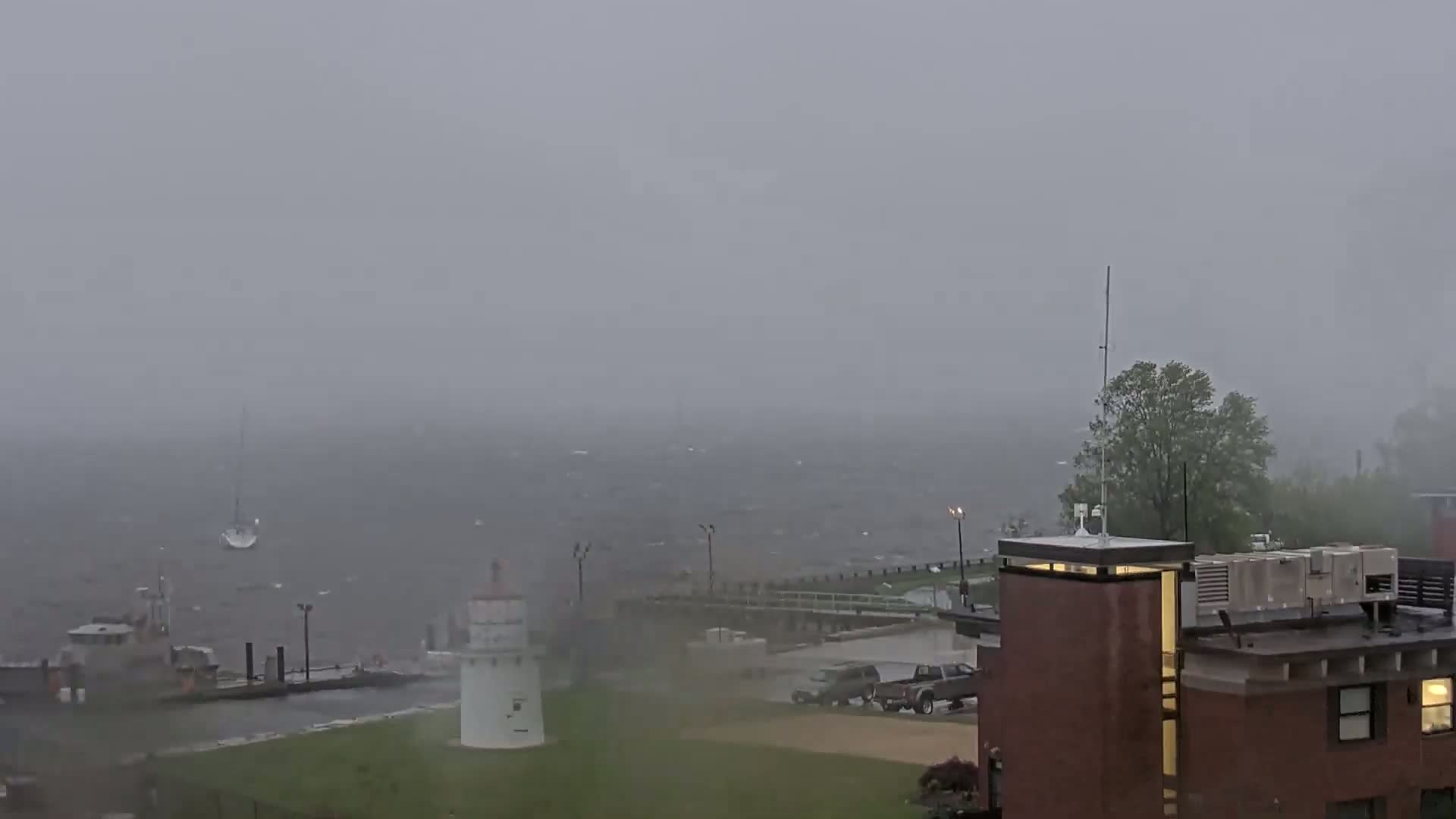 A hazy, overcast day shows a small sailboat on choppy water, near a lighthouse and a brick building, with some vehicles parked nearby.