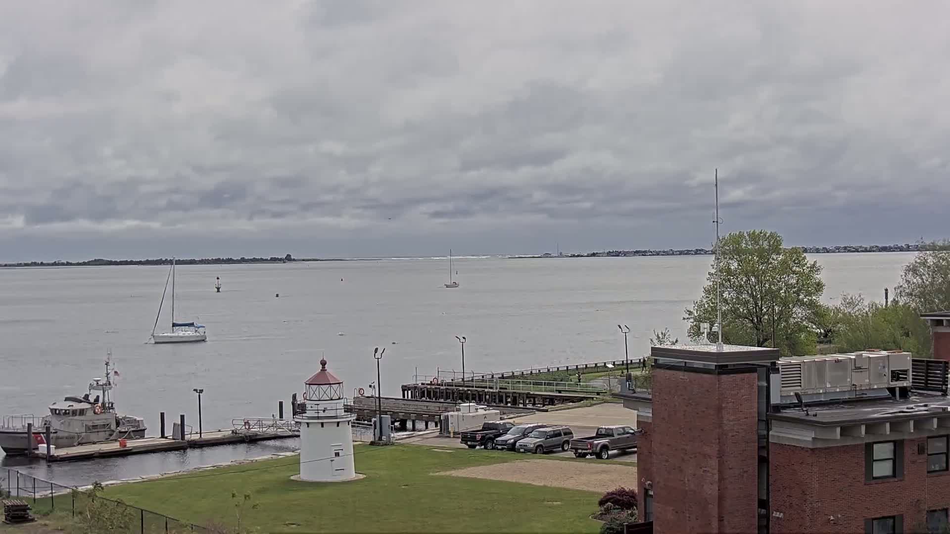 A small lighthouse and several boats are visible near a waterfront building under an overcast sky.