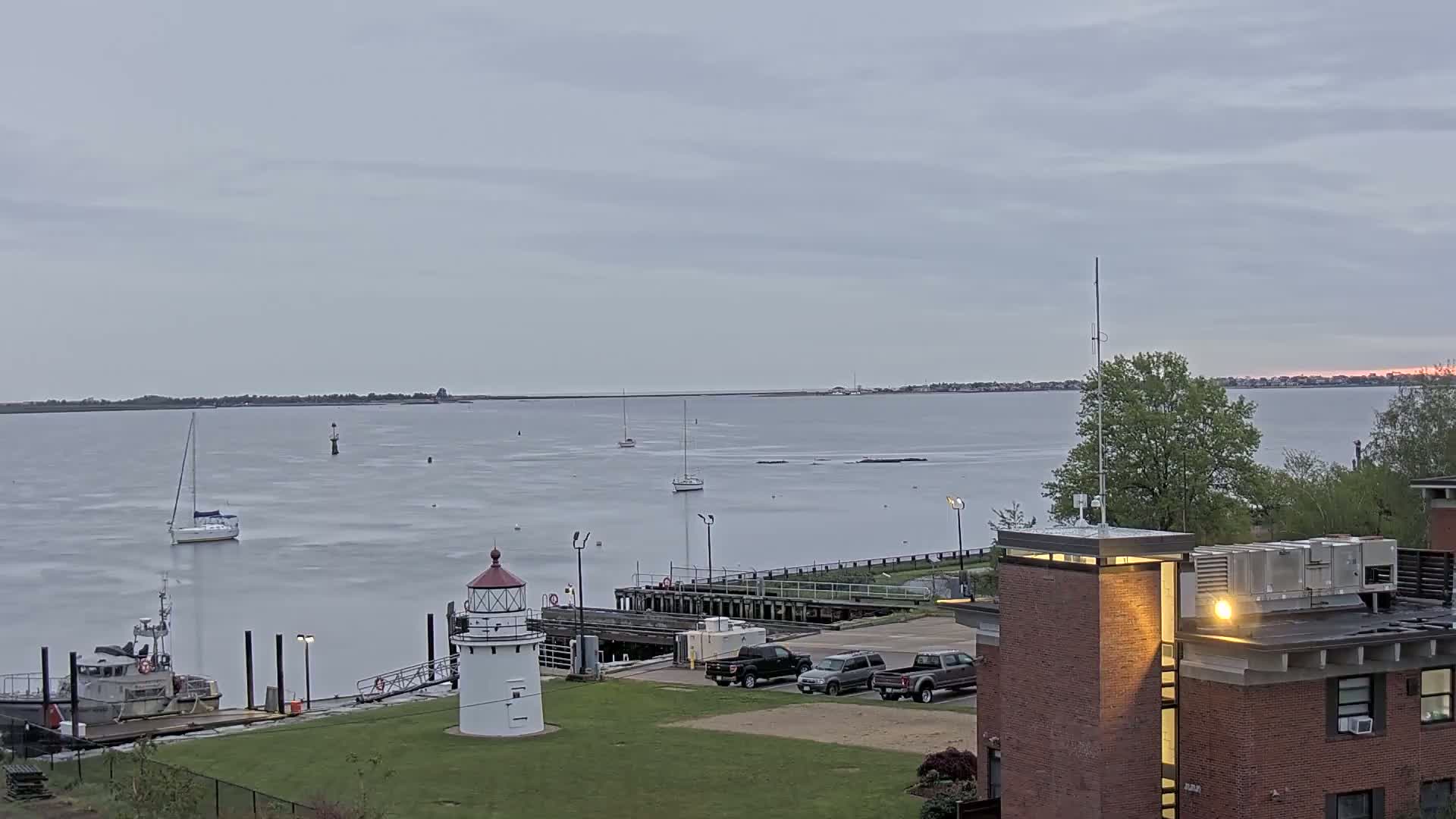 A small lighthouse sits on a grassy area overlooking a calm body of water with several sailboats and a small ship, under an overcast sky.