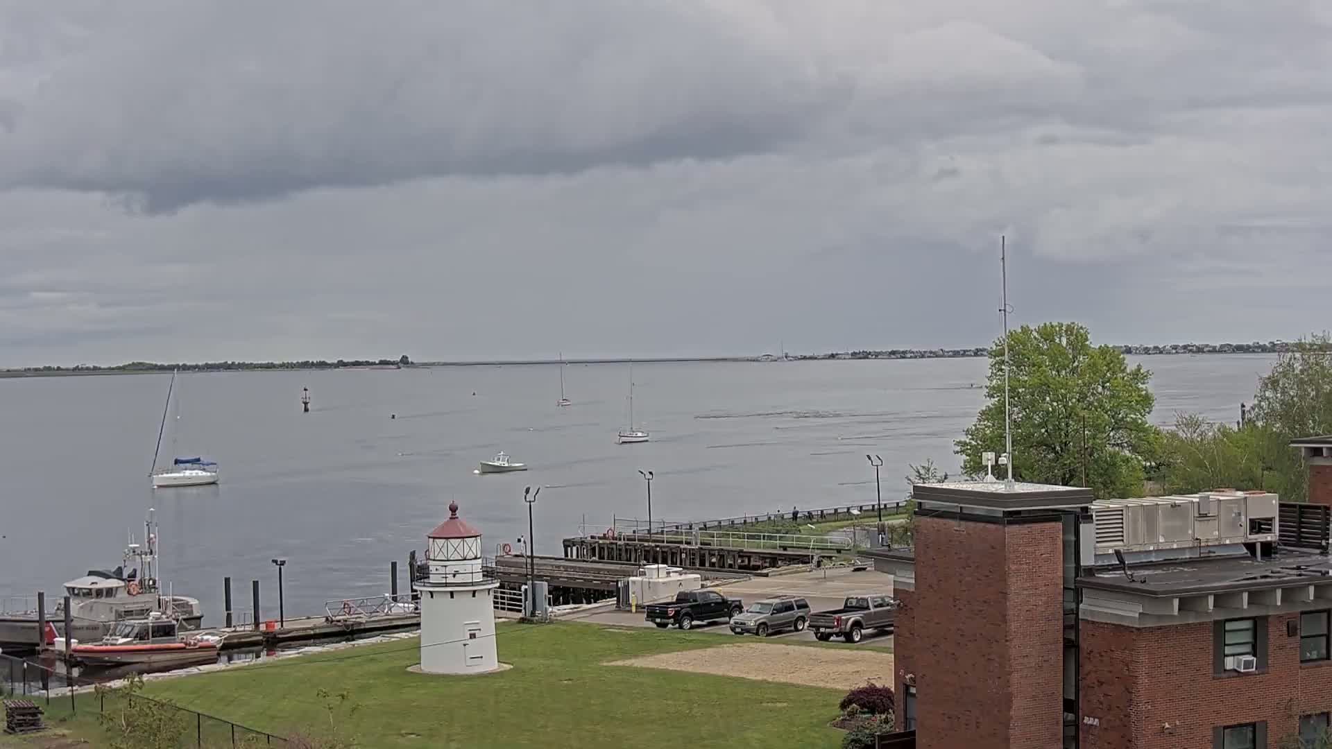 A calm, overcast day shows a harbor with several boats, a lighthouse, and a Coast Guard vessel moored at a dock near a brick building.