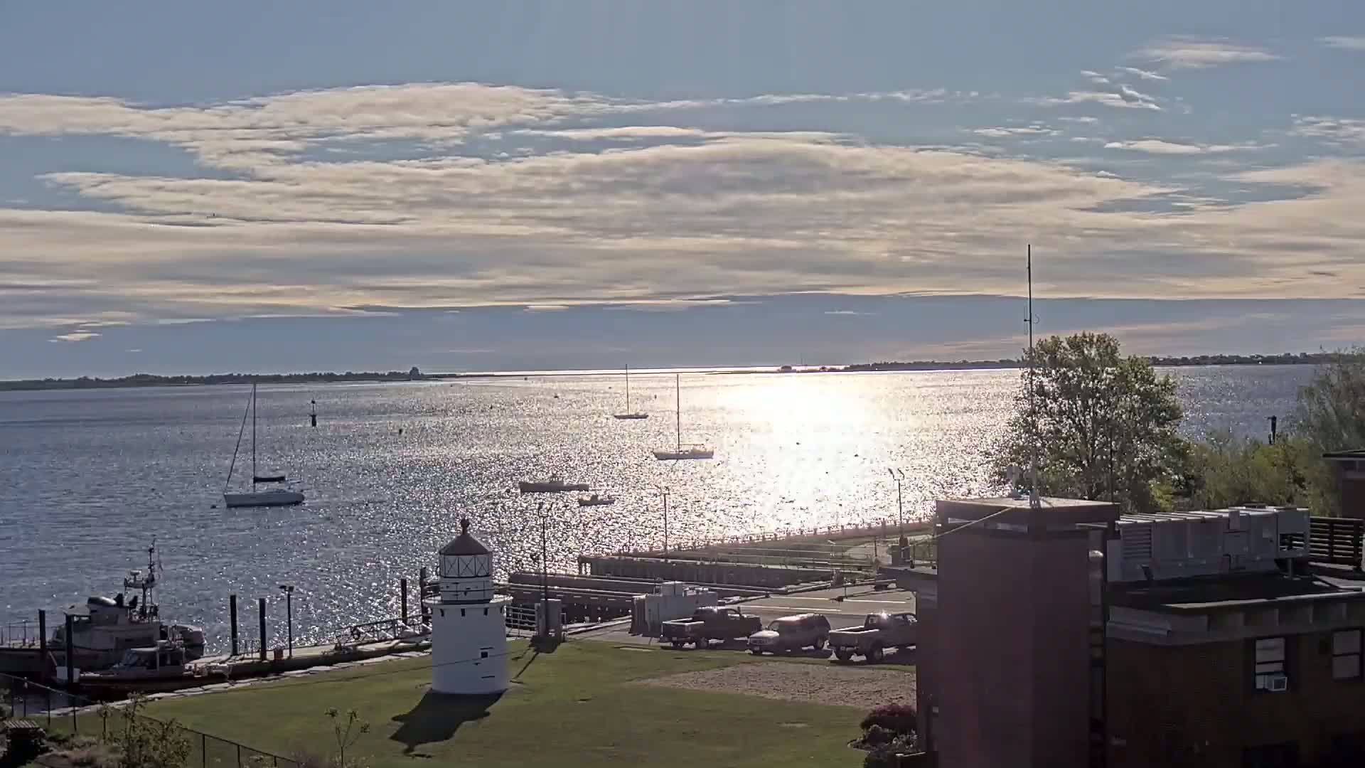 A sparkling body of water with several sailboats is viewed from a waterfront location under a partly cloudy sky, showcasing a small lighthouse and a tugboat near the shore.