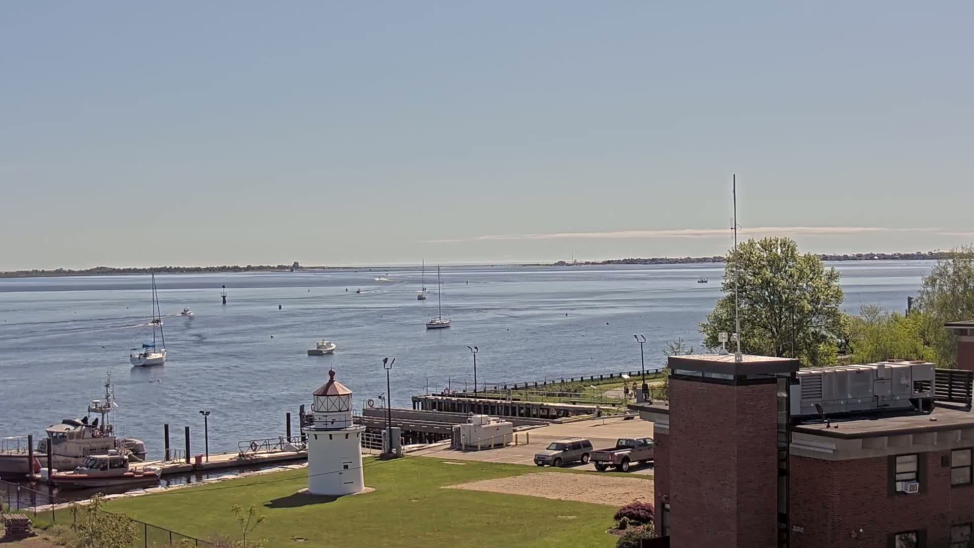 A calm body of water with several sailboats and motorboats, a small lighthouse, a Coast Guard vessel at a dock, and a brick building on a sunny day.