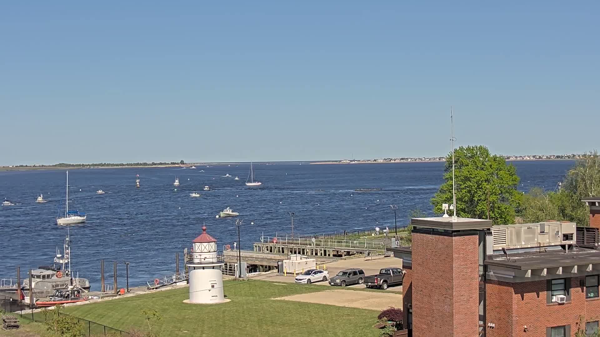 A coastal scene on a sunny day shows a body of water with numerous boats, a lighthouse, a Coast Guard vessel, and several cars parked in a lot near a brick building.