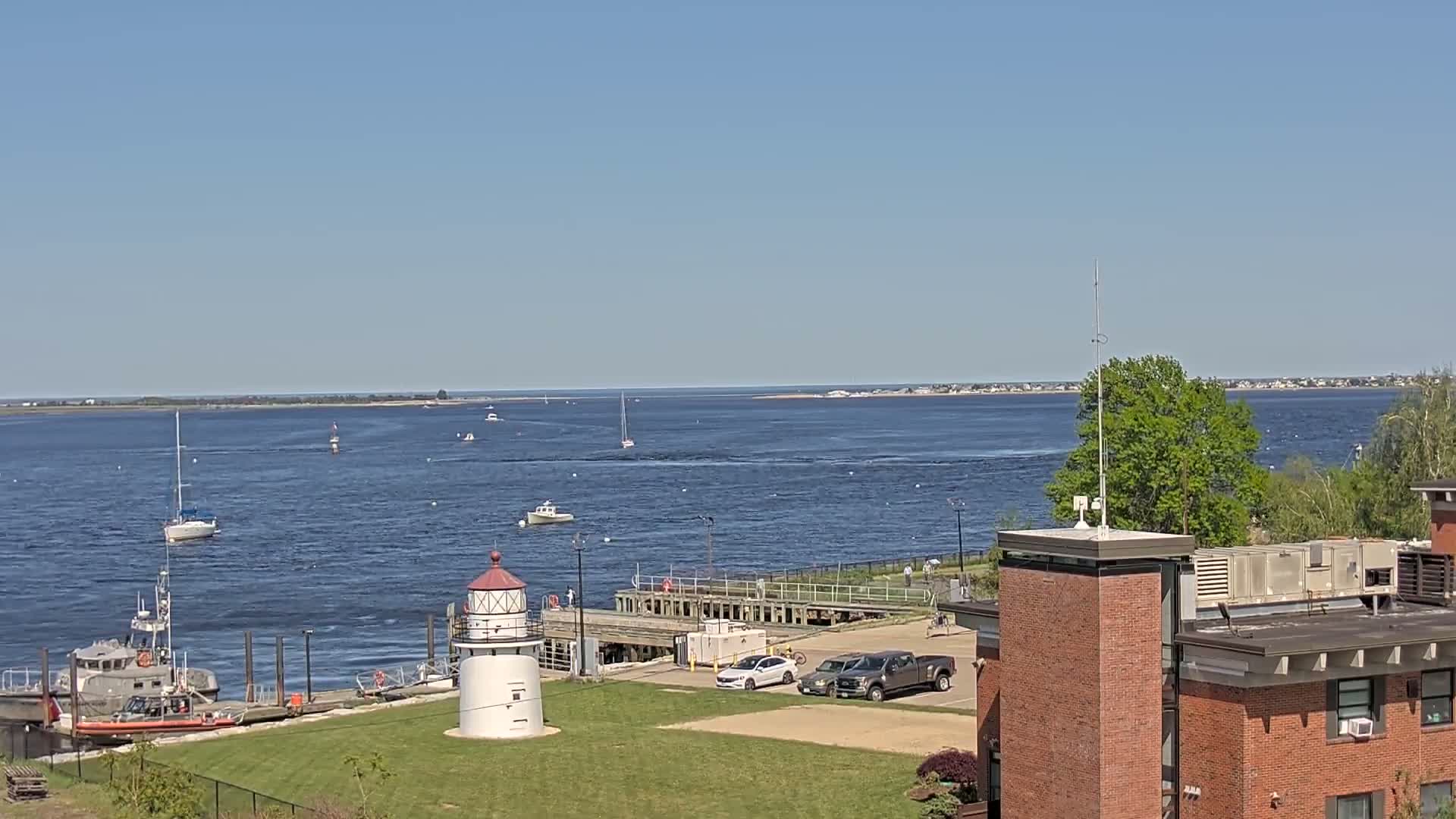 A small lighthouse sits on a grassy area next to a body of water with several boats and a Coast Guard vessel, under a clear blue sky.
