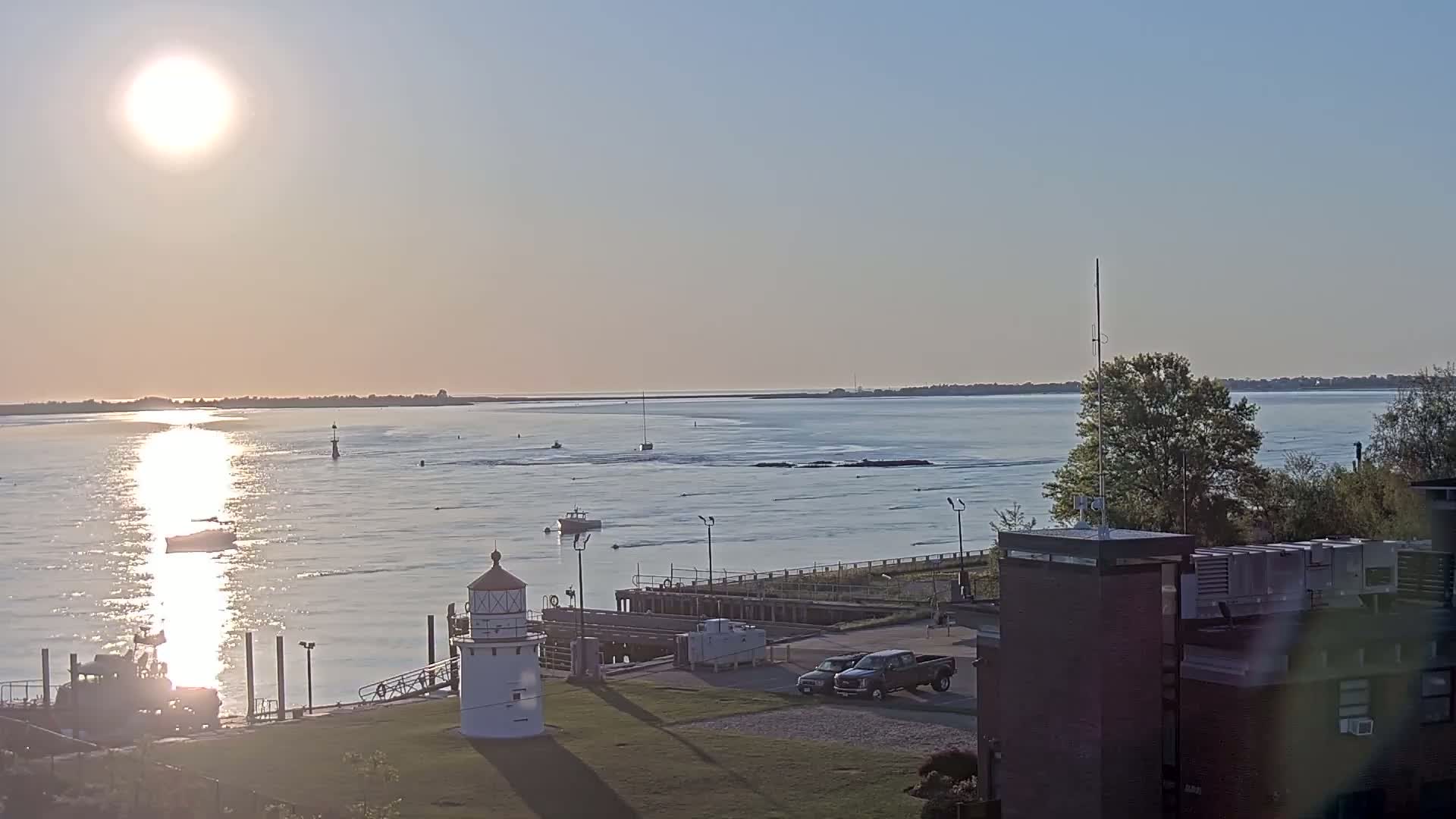 A calm body of water under a bright sunrise shows several boats, a lighthouse, and a building with a grassy area in the foreground.