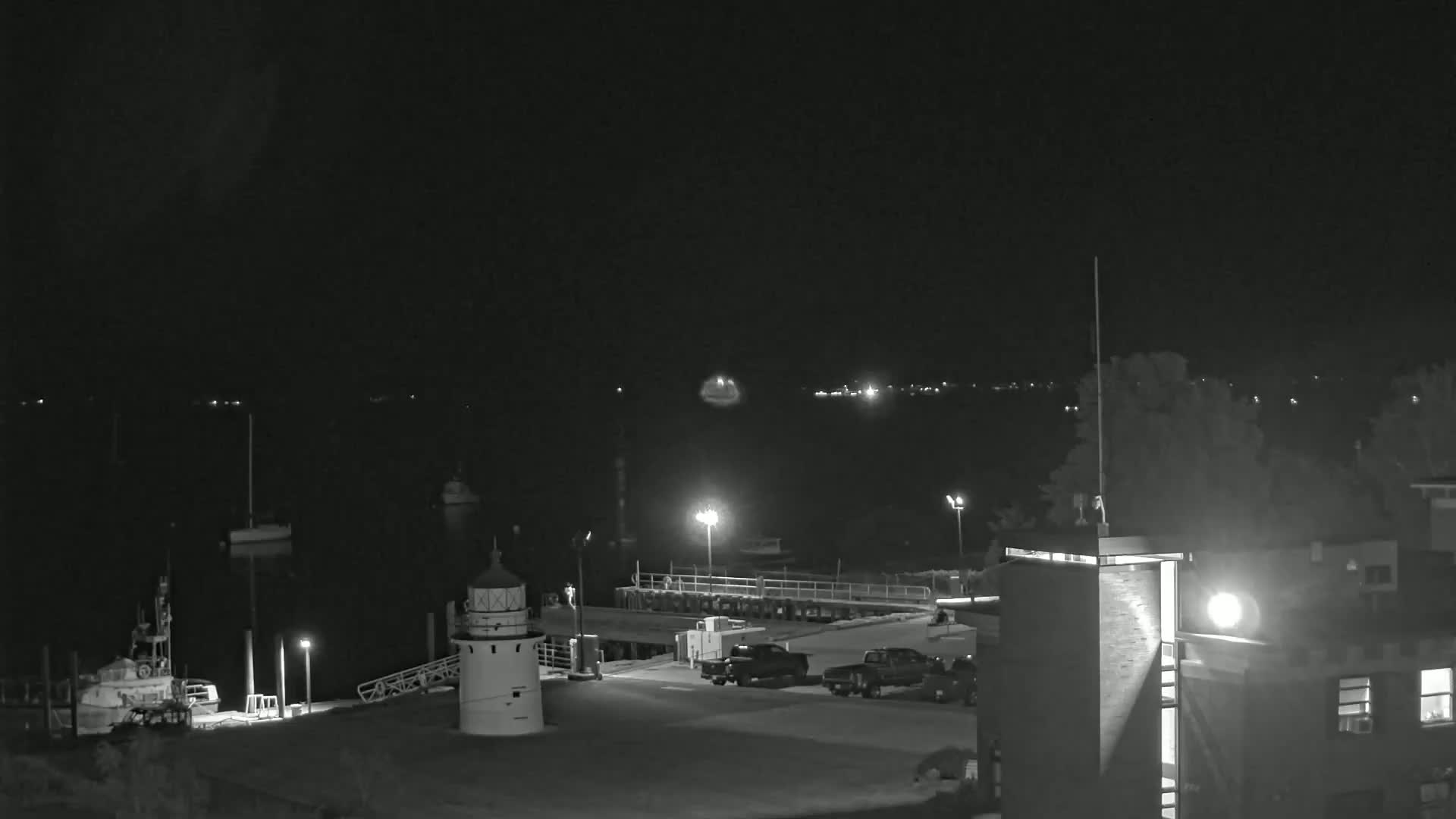 A nighttime, low-light view of a harbor with a lighthouse, boats, and vehicles parked in a lot next to a building.