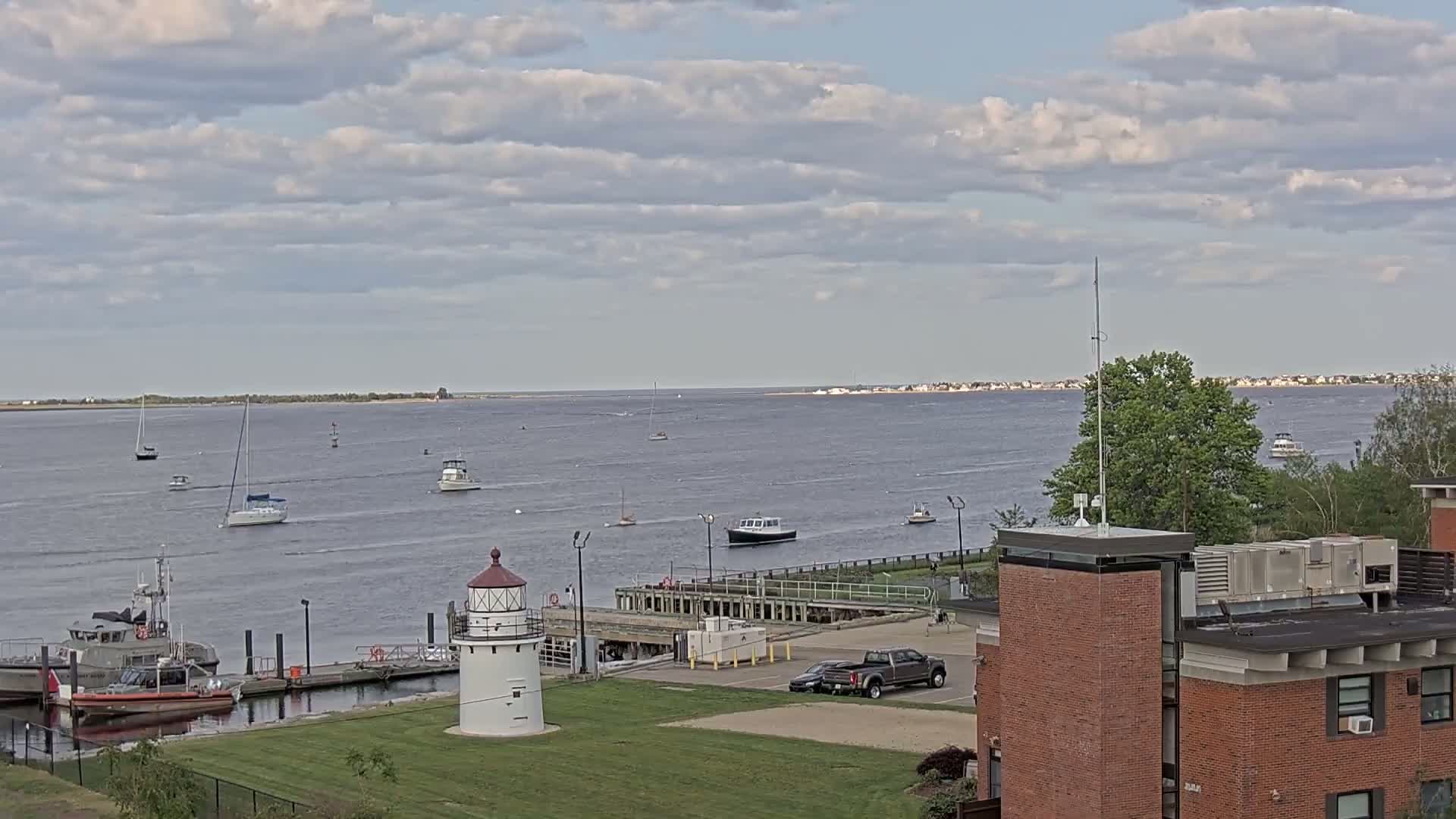 A harbor scene under a partly cloudy sky shows several boats on the water, a lighthouse, Coast Guard vessels at a dock, and a brick building on the shore.