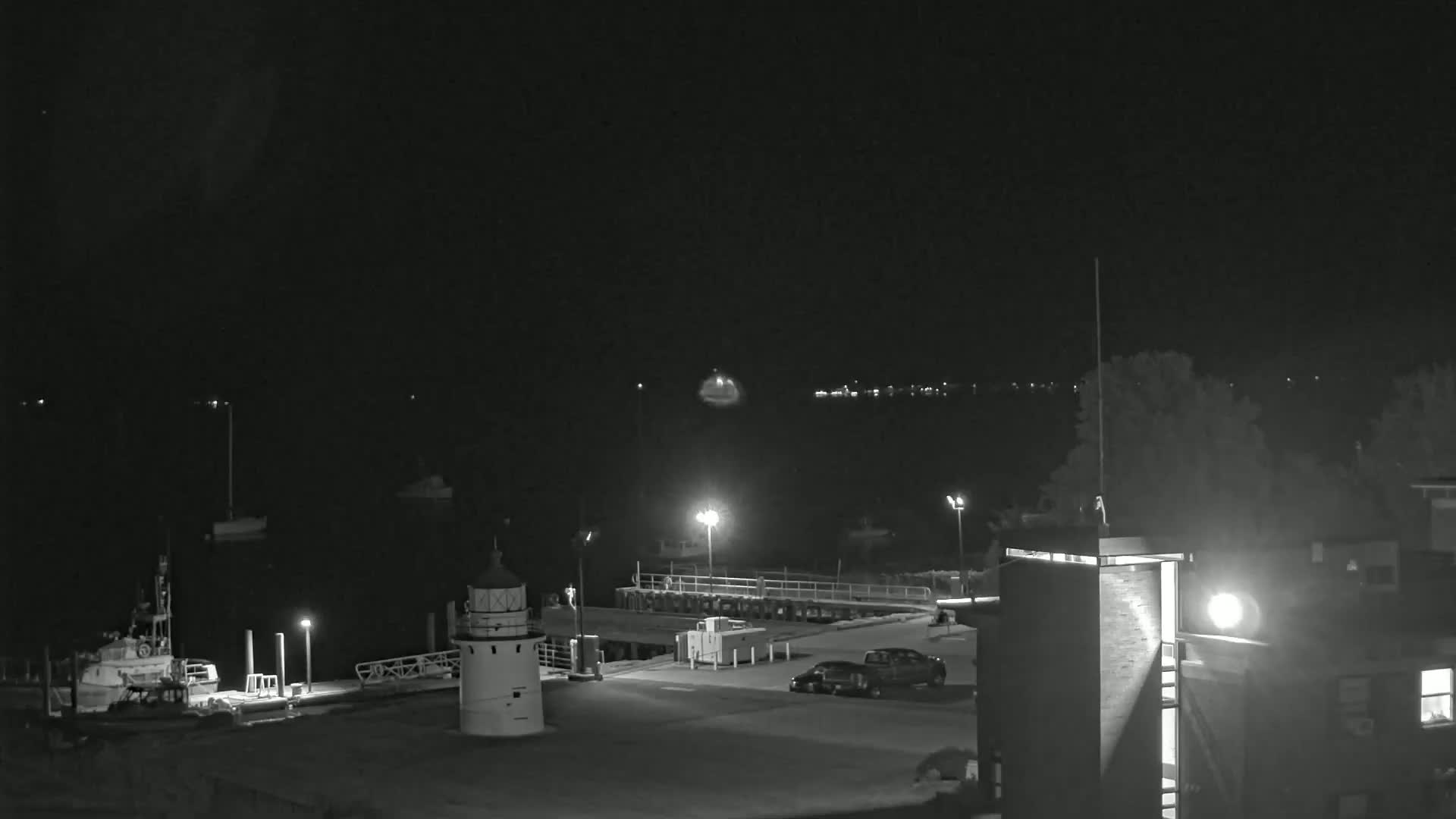 A nighttime view of a harbor with a lighthouse, boats, a dock, and a few vehicles parked in a lot, under a clear, dark sky.