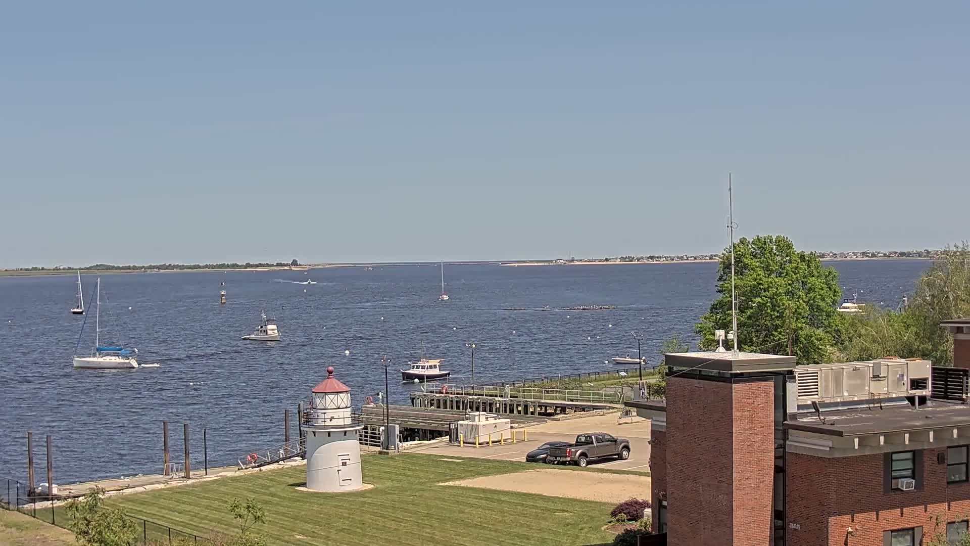 A calm, sunny day shows a body of water with sailboats and motorboats, a lighthouse on a grassy area near a brick building, and a distant shoreline.