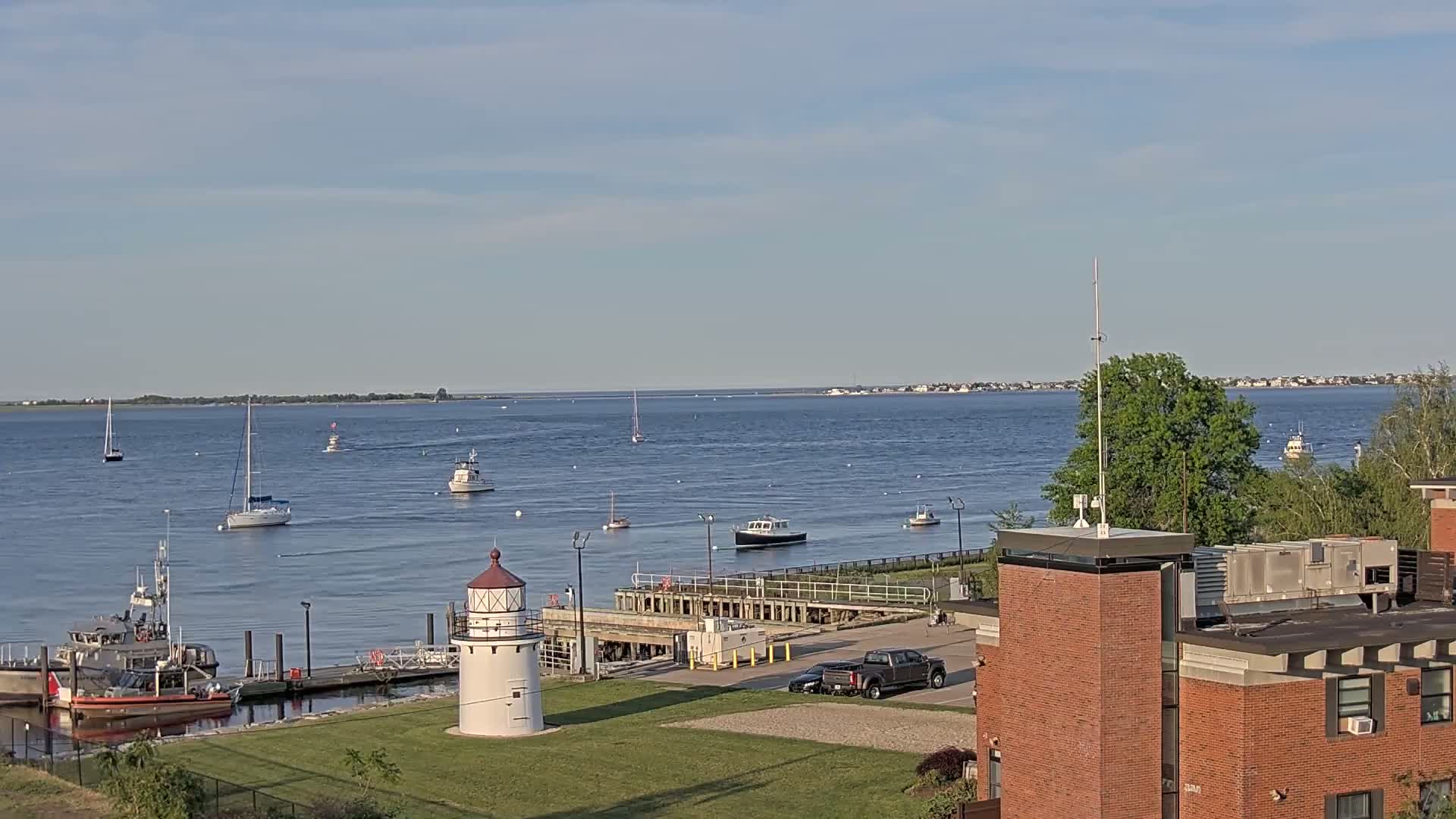 A calm body of water with several sailboats and motorboats, a lighthouse, and a brick building are visible under a mostly clear sky.