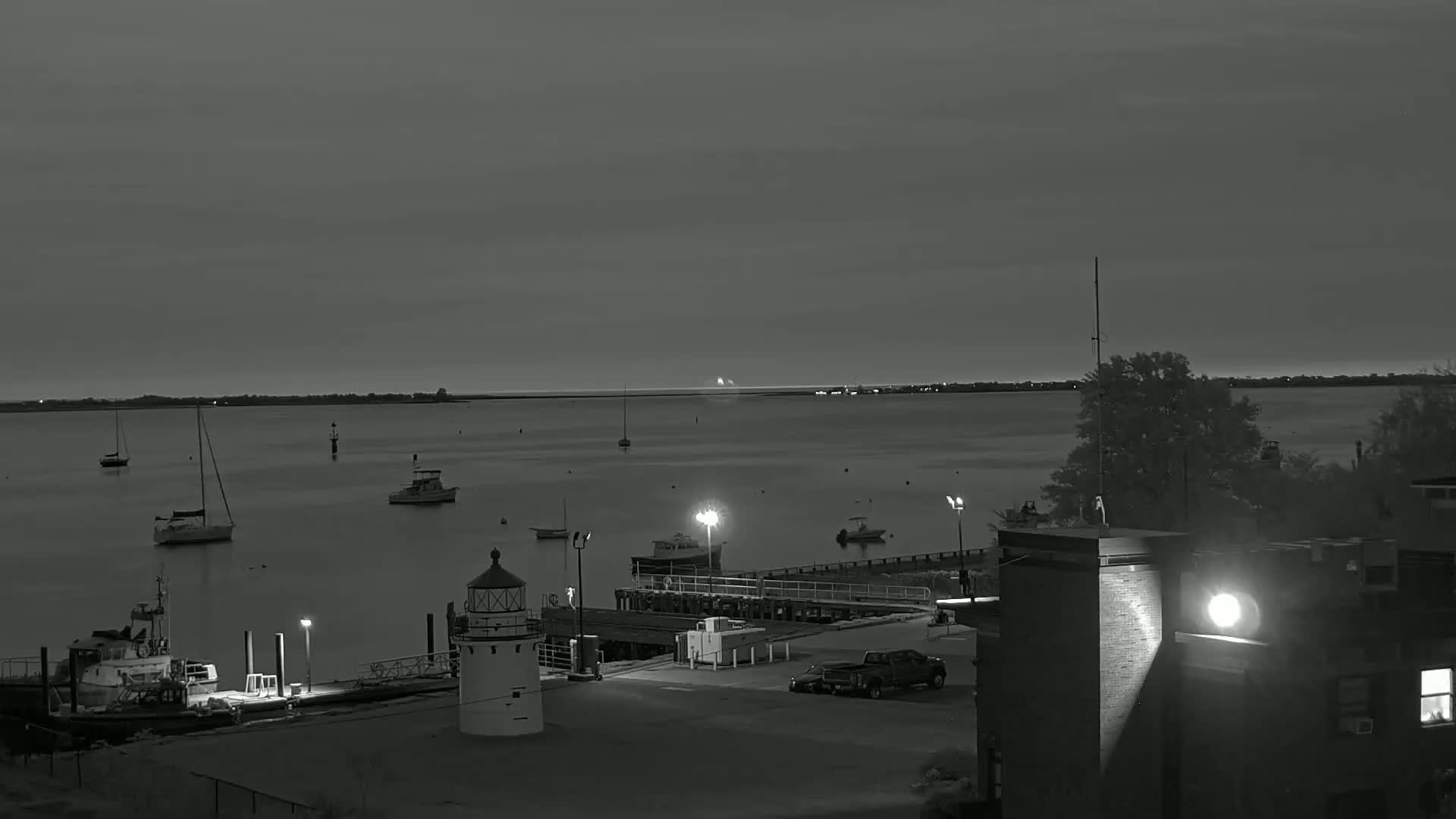 A nighttime, black-and-white view of a harbor with several boats moored near a lighthouse and a building, under a mostly clear sky.