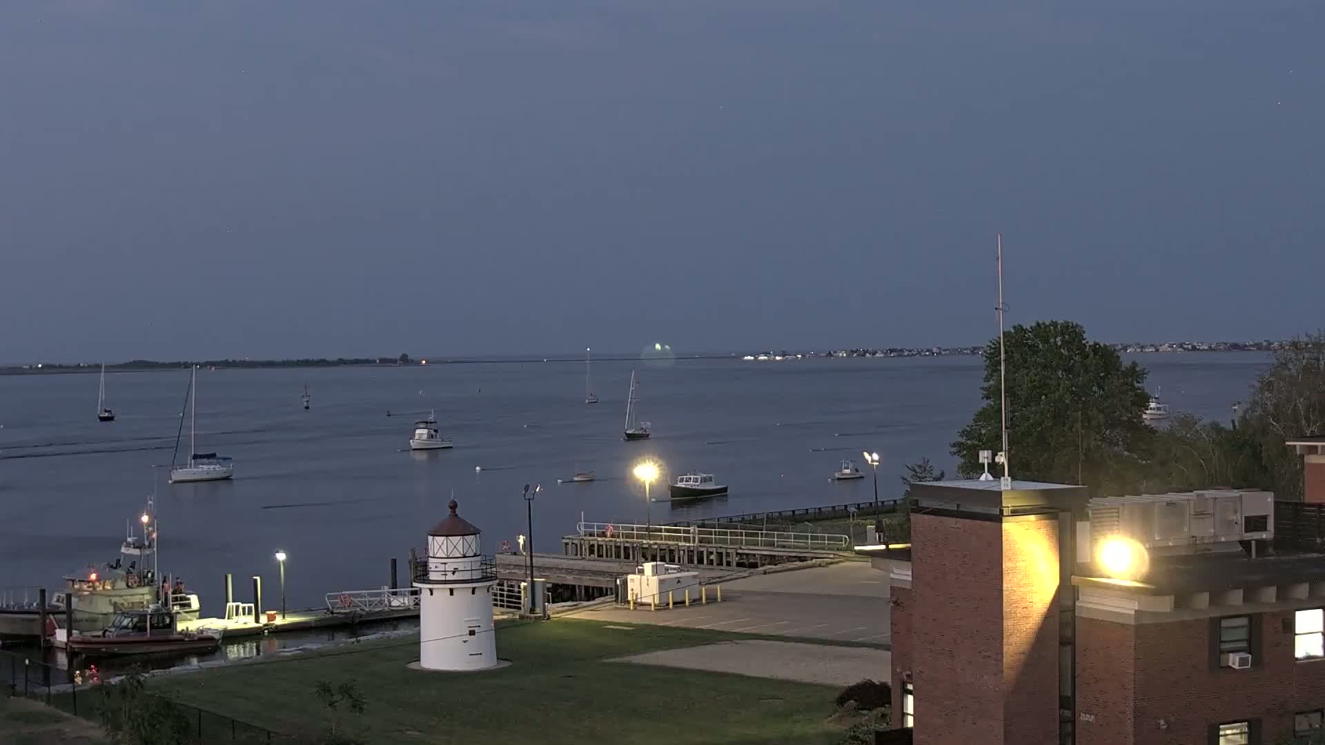 A small lighthouse stands on a grassy area next to a dock with several boats and a larger vessel at twilight under a clear, dusky sky.