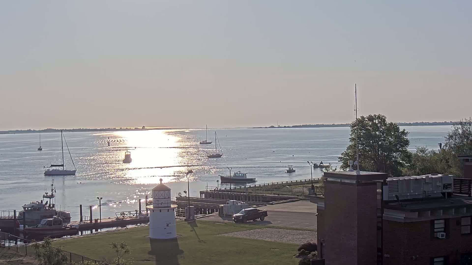 A calm, sunny day shows a harbor with several boats, a lighthouse, and a building on the shore.