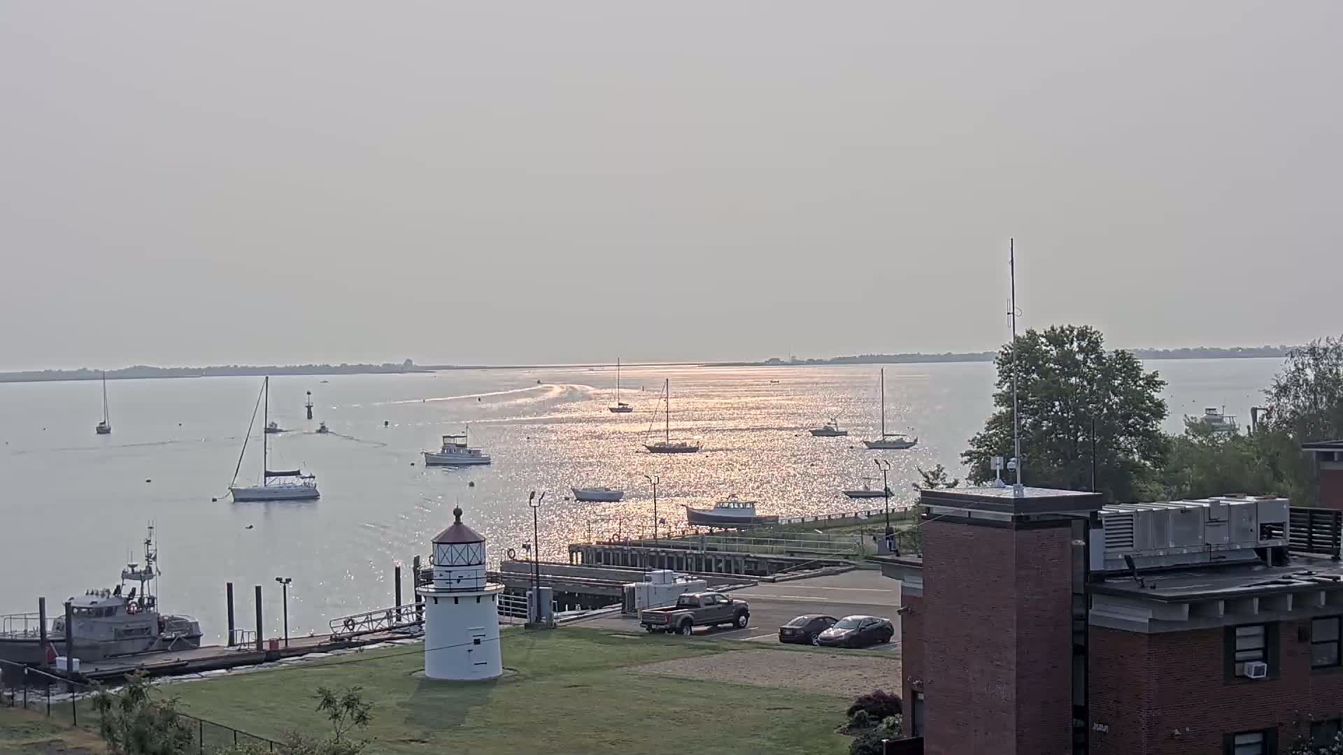 A hazy, sunny day reveals a calm harbor with numerous sailboats and motorboats, a lighthouse, and a small dock, observed from an elevated vantage point near a building.