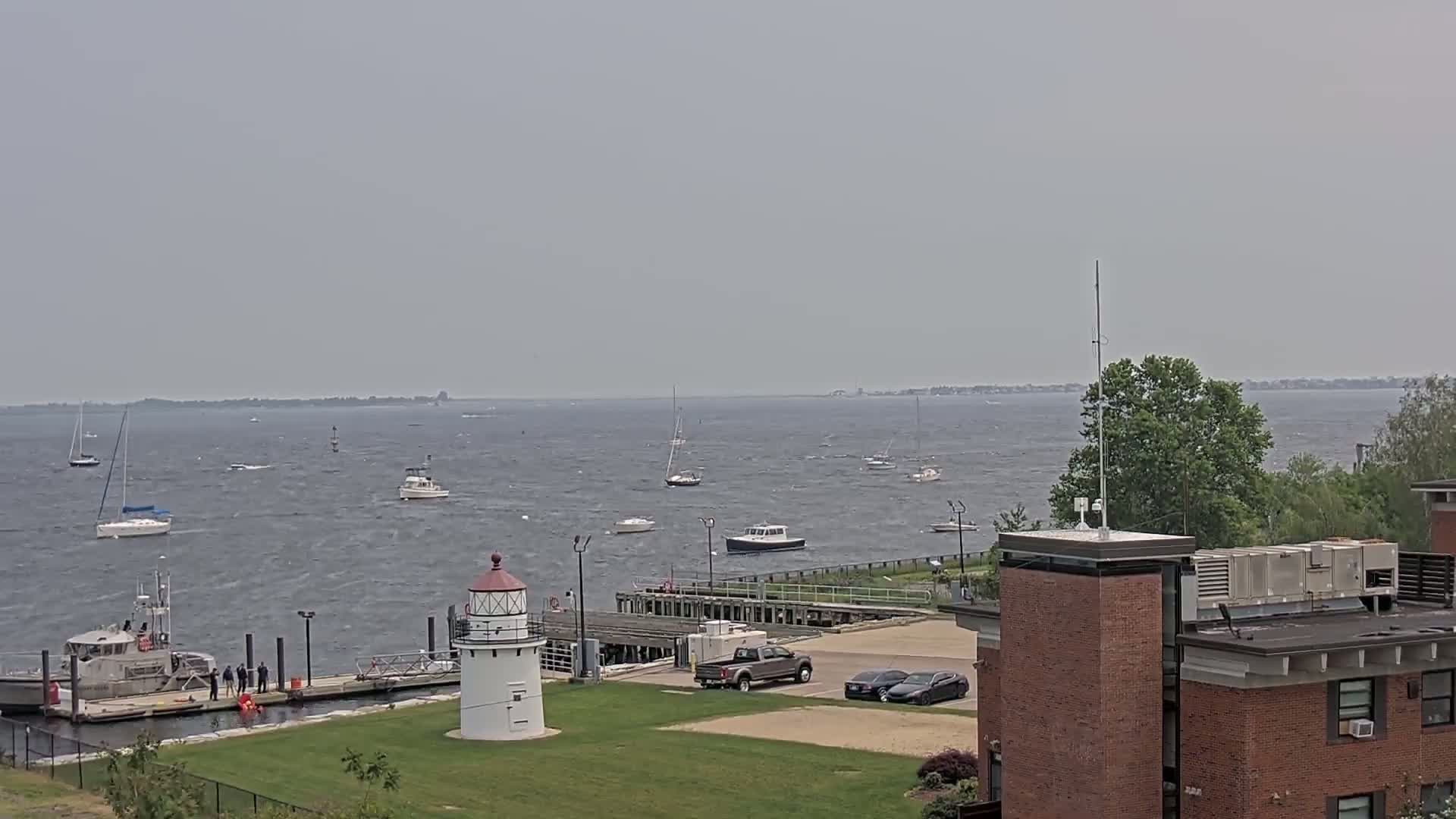 A body of water with several sailboats and motorboats under a hazy sky, a small lighthouse, and a dock with a small ship and people are visible near a brick building.