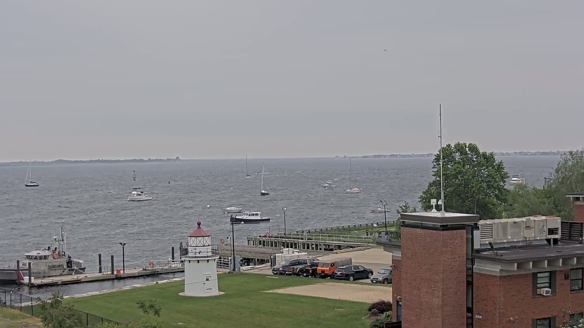 A lighthouse stands on a grassy area next to a harbor with several boats and a Coast Guard vessel, under an overcast sky.