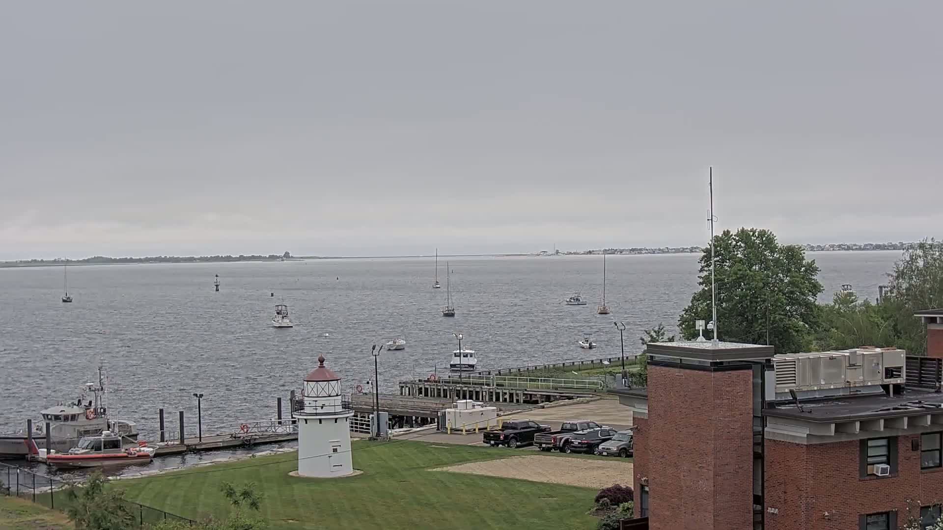 A Coast Guard vessel is docked near a small lighthouse, overlooking a calm body of water with several sailboats and other small watercraft under an overcast sky.