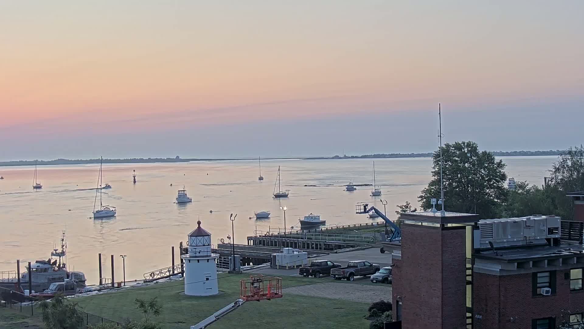 A calm body of water with numerous sailboats and a small lighthouse at sunset, next to a dock with several vehicles and buildings.