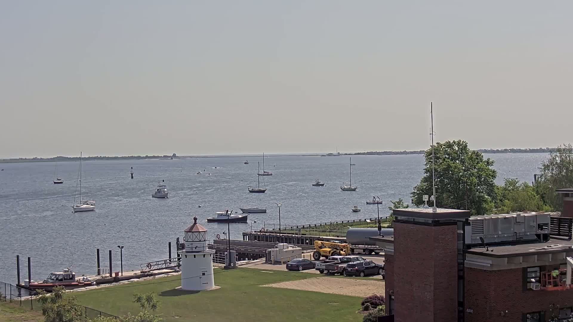 A calm, sunny day reveals a harbor scene with several sailboats and motorboats, a lighthouse on a grassy area next to a dock, and a building in the foreground.
