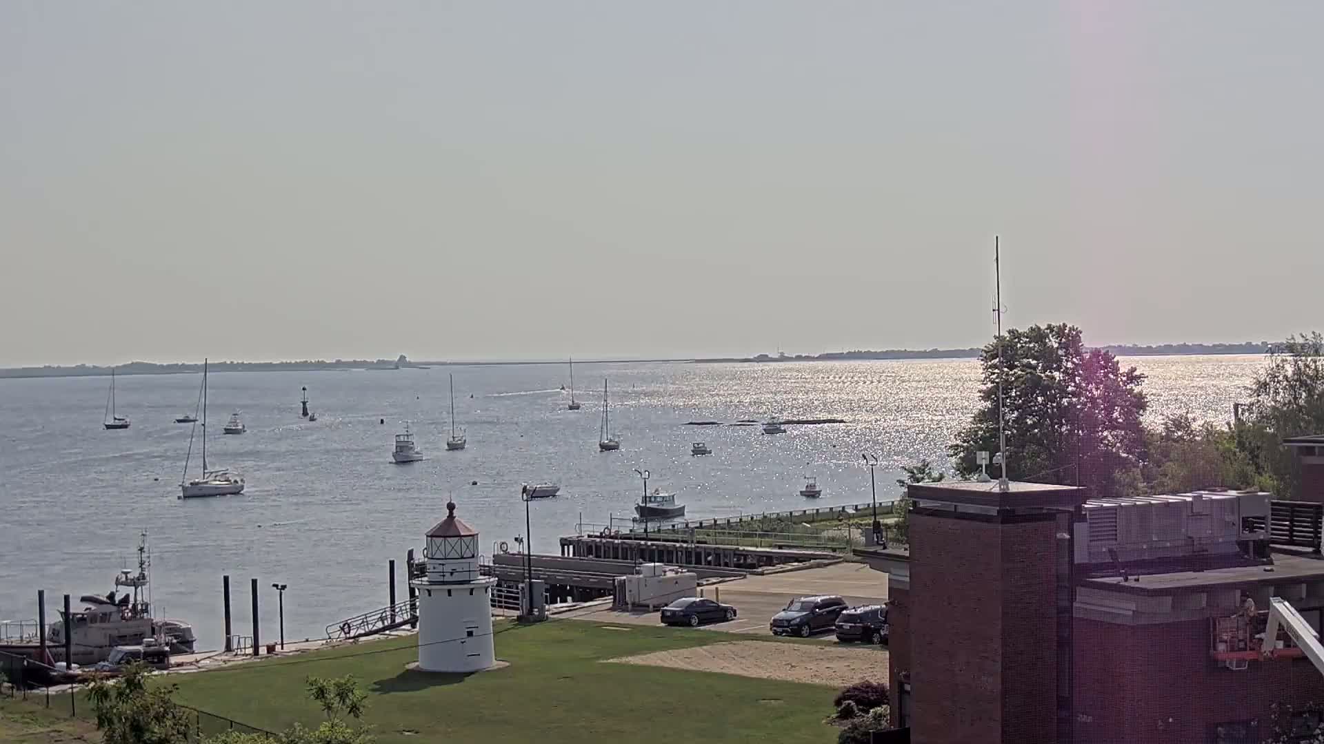 A calm, sunny day shows a harbor with numerous sailboats and motorboats, a small lighthouse, and a brick building in the foreground.