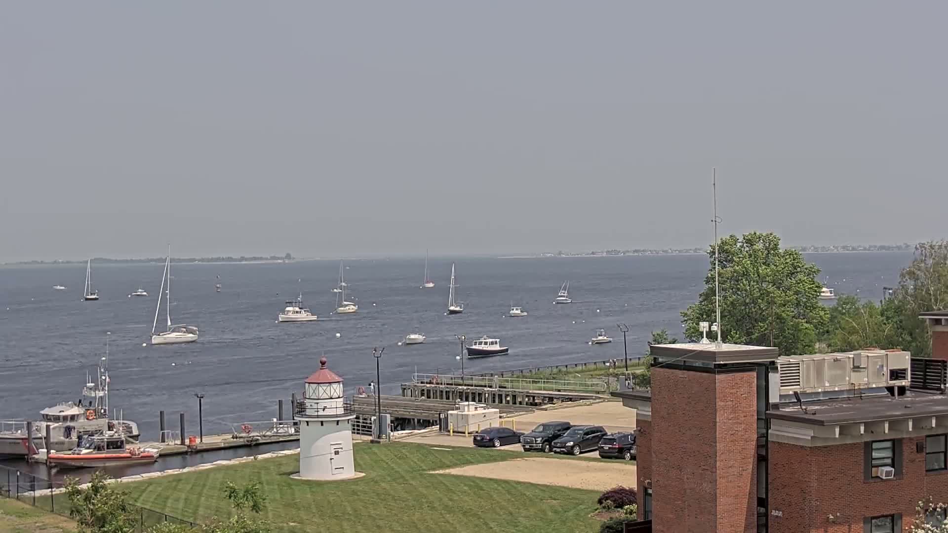 A hazy day shows several sailboats and motorboats on the water, a lighthouse on the shore near a Coast Guard station, and a brick building in the foreground.