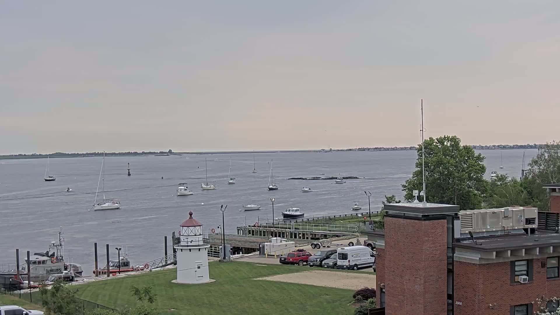 A lighthouse stands on a grassy area near a harbor filled with sailboats and motorboats under an overcast sky.
