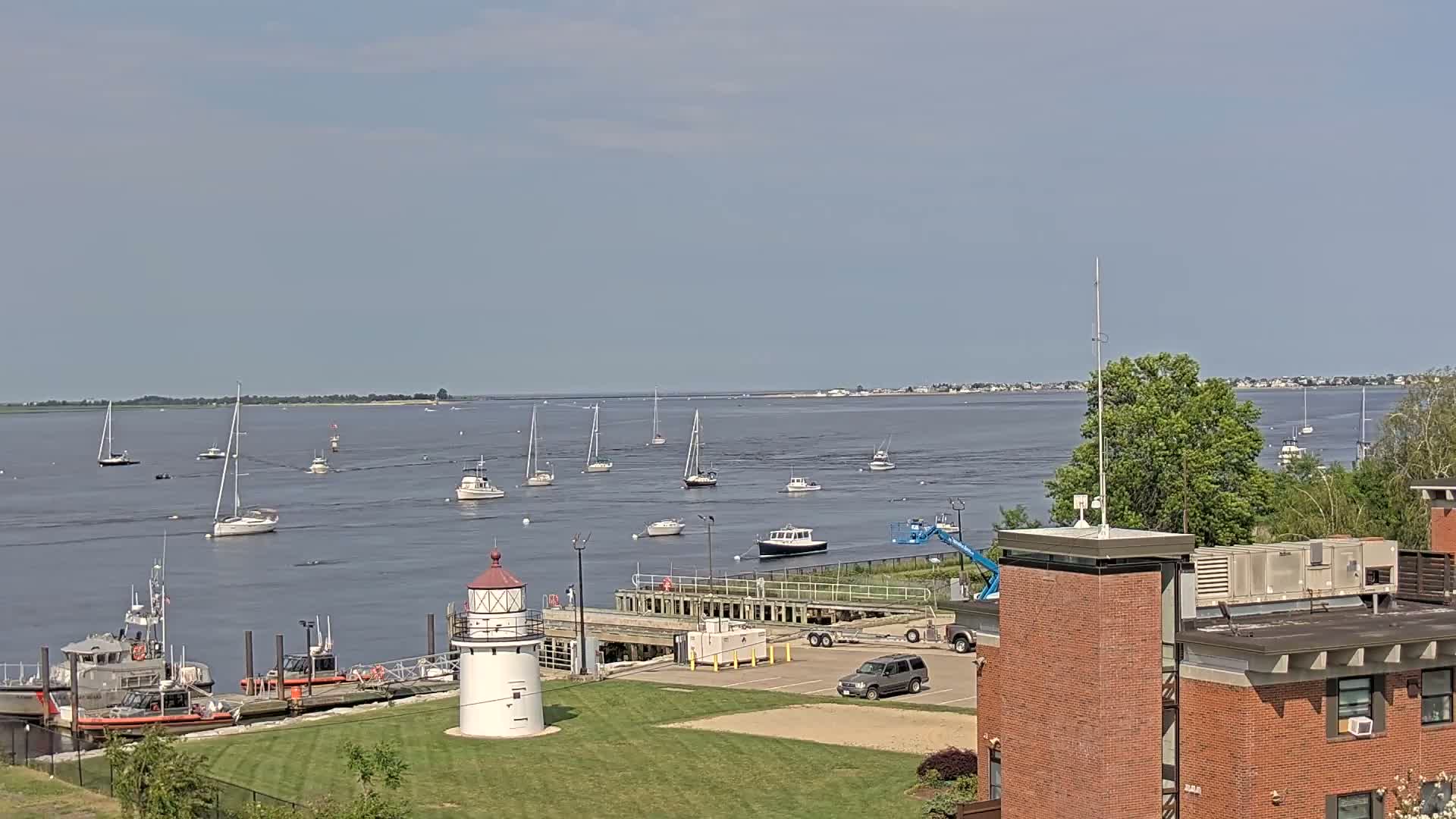 A calm, sunny day reveals a harbor scene with numerous sailboats and motorboats, a lighthouse, Coast Guard vessels, and a brick building.