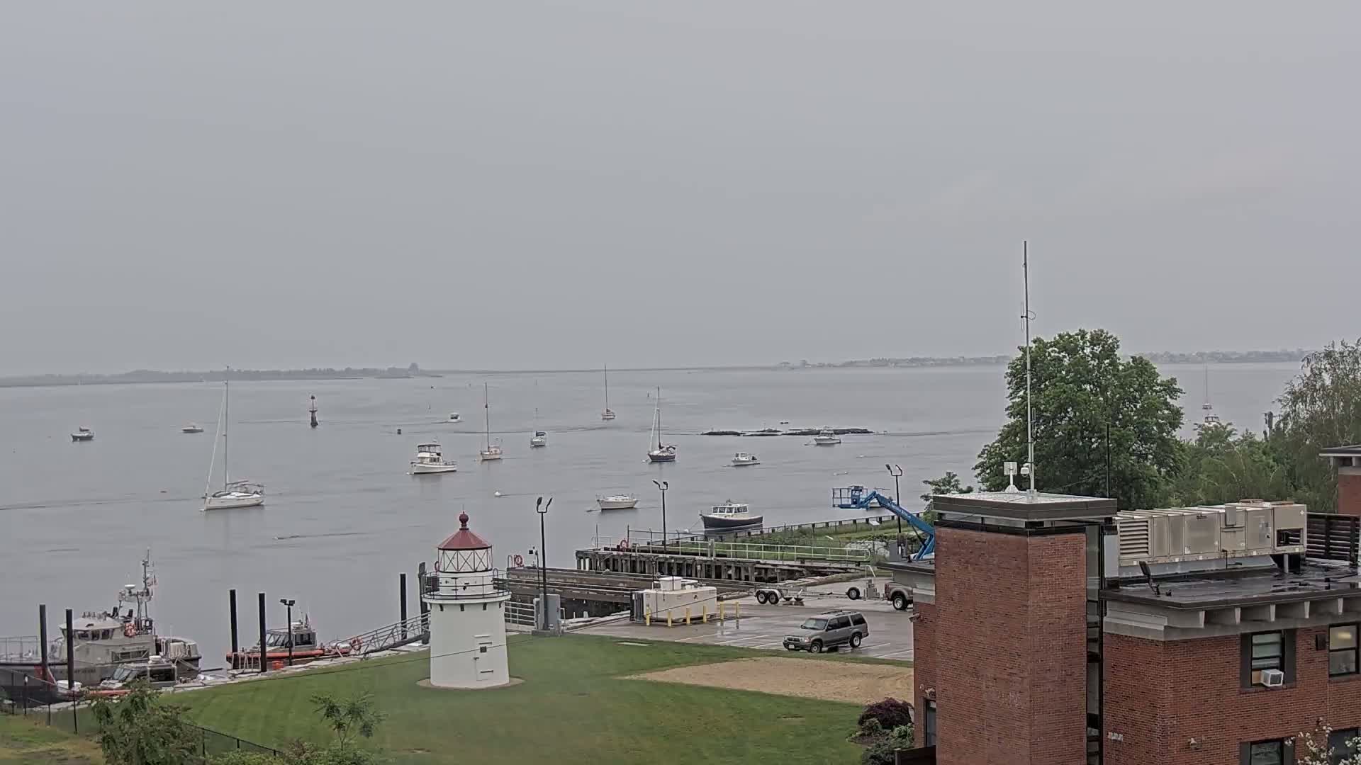 A harbor scene on an overcast day shows several boats and a lighthouse near a brick building.