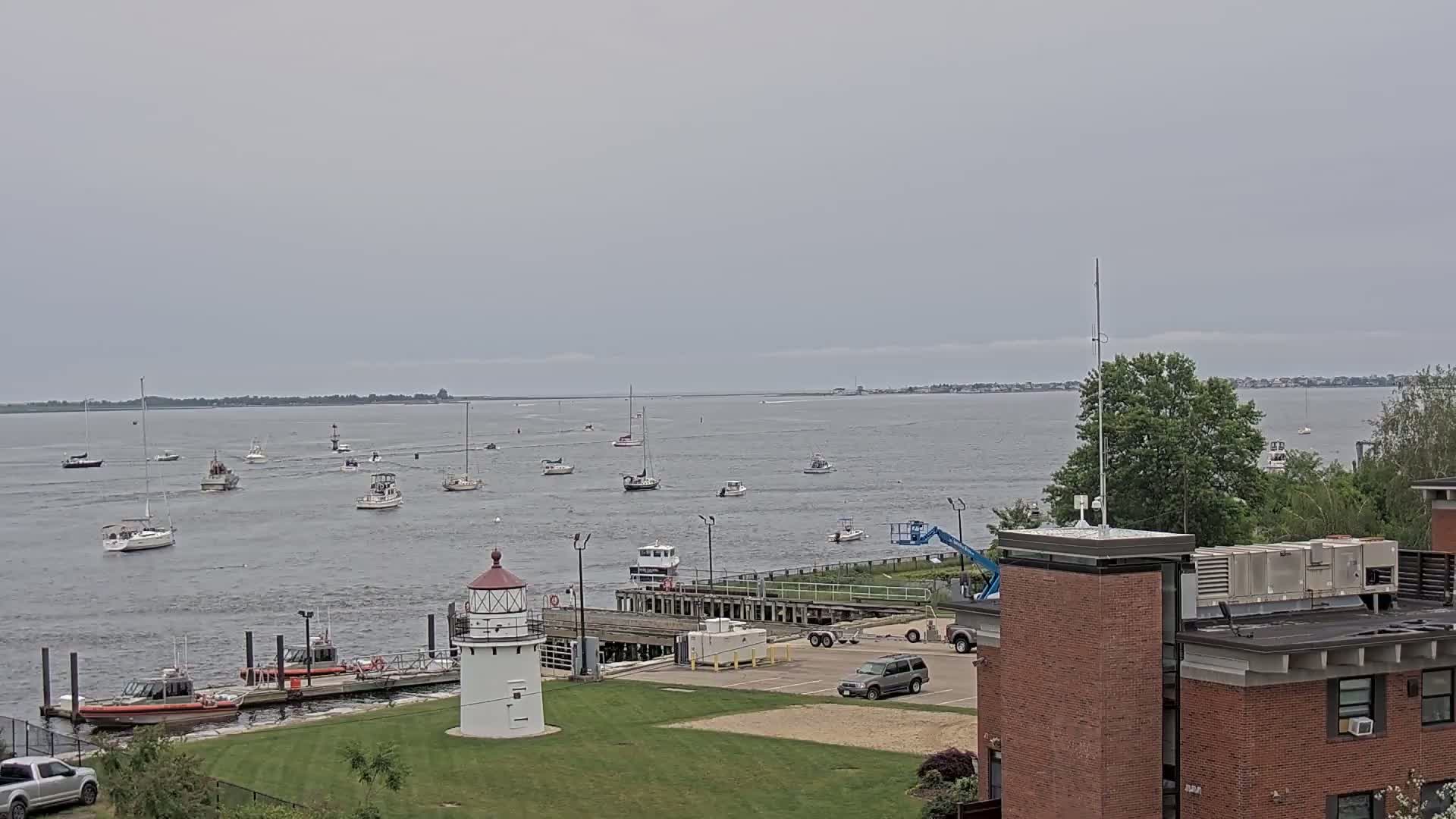 Numerous boats are scattered across a calm body of water under an overcast sky, with a lighthouse and a brick building visible in the foreground.