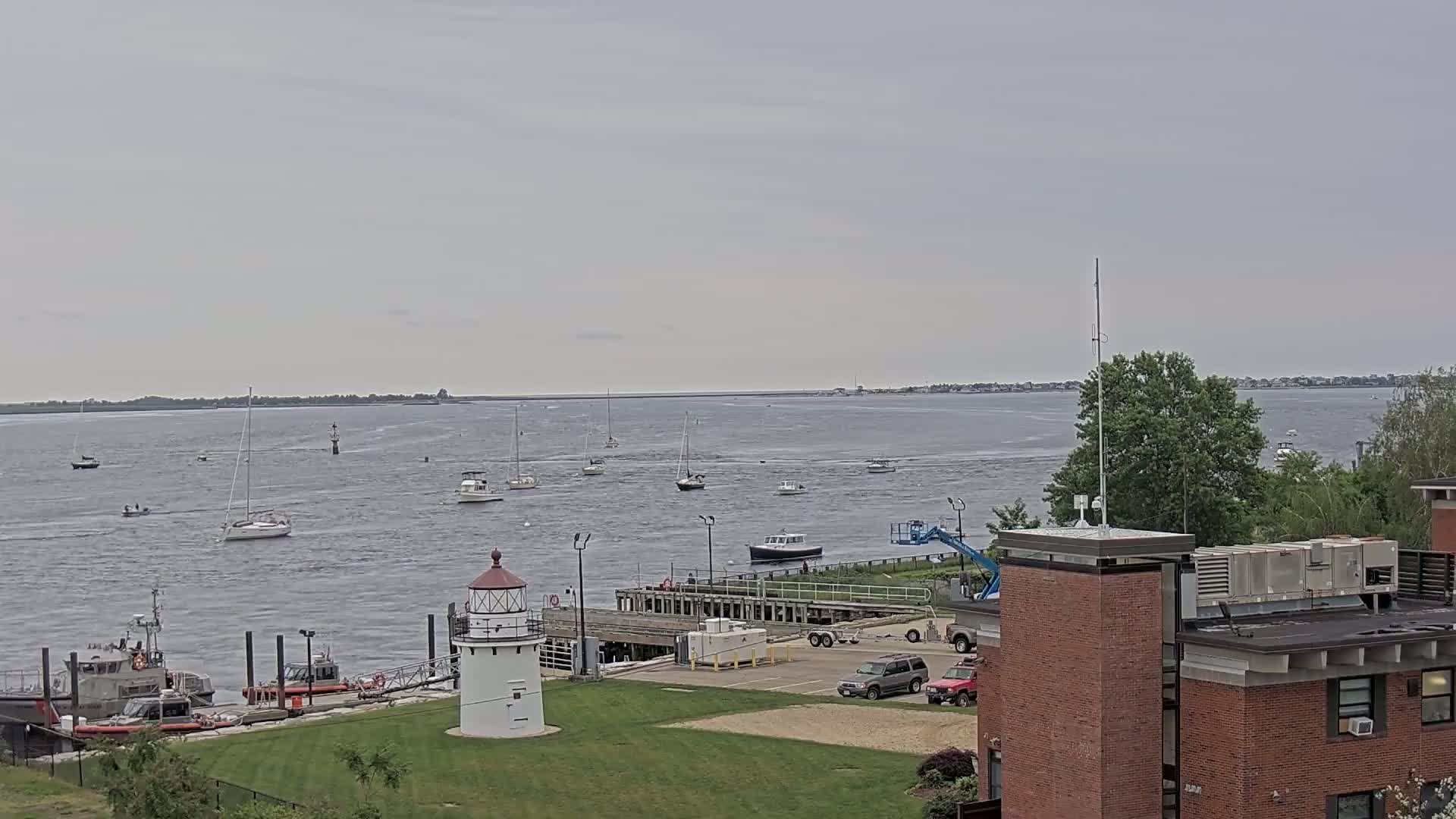Numerous boats are scattered across a calm body of water under an overcast sky, with a lighthouse and a brick building visible in the foreground.