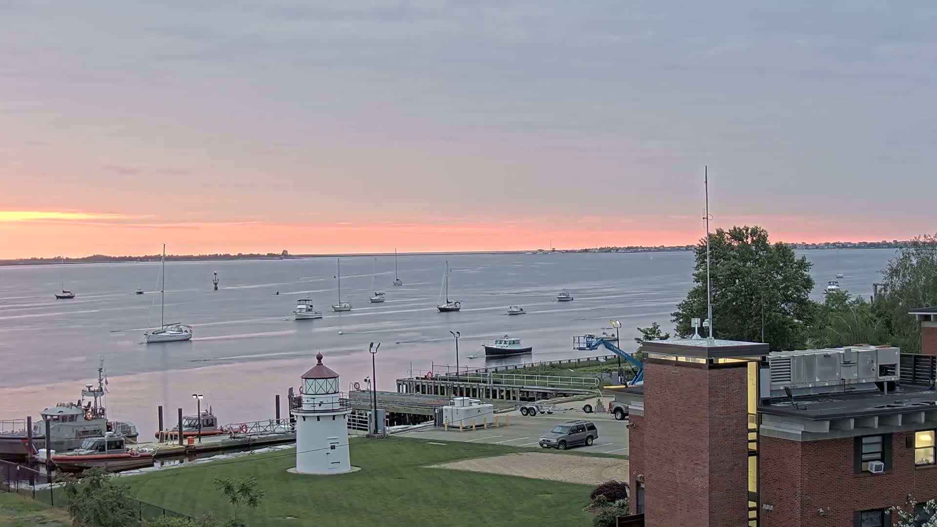 A pink sunset over a calm body of water with numerous sailboats and motorboats, a lighthouse, and Coast Guard vessels docked near a brick building.
