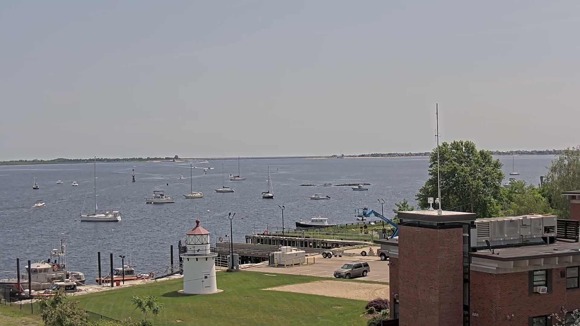 A calm, sunny day reveals a harbor filled with sailboats and motorboats, a small lighthouse, and a Coast Guard vessel near a brick building.