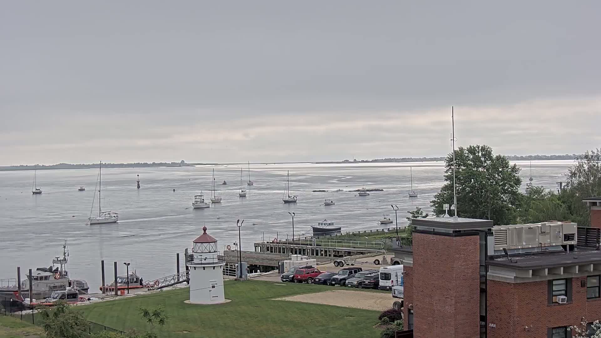 A calm, overcast day shows a harbor with numerous sailboats and a lighthouse near a brick building, with a Coast Guard vessel moored nearby.
