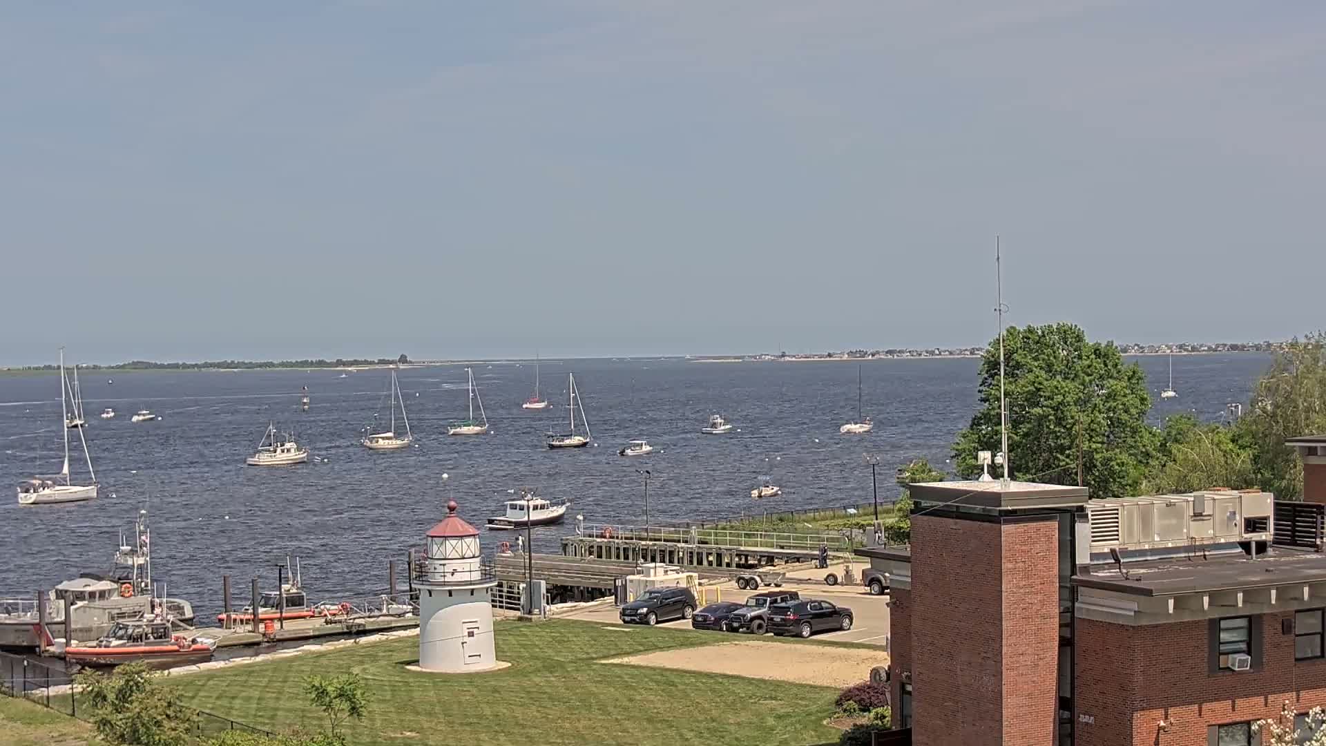 A small lighthouse stands on a grassy area overlooking a harbor filled with sailboats and Coast Guard vessels on a sunny day.