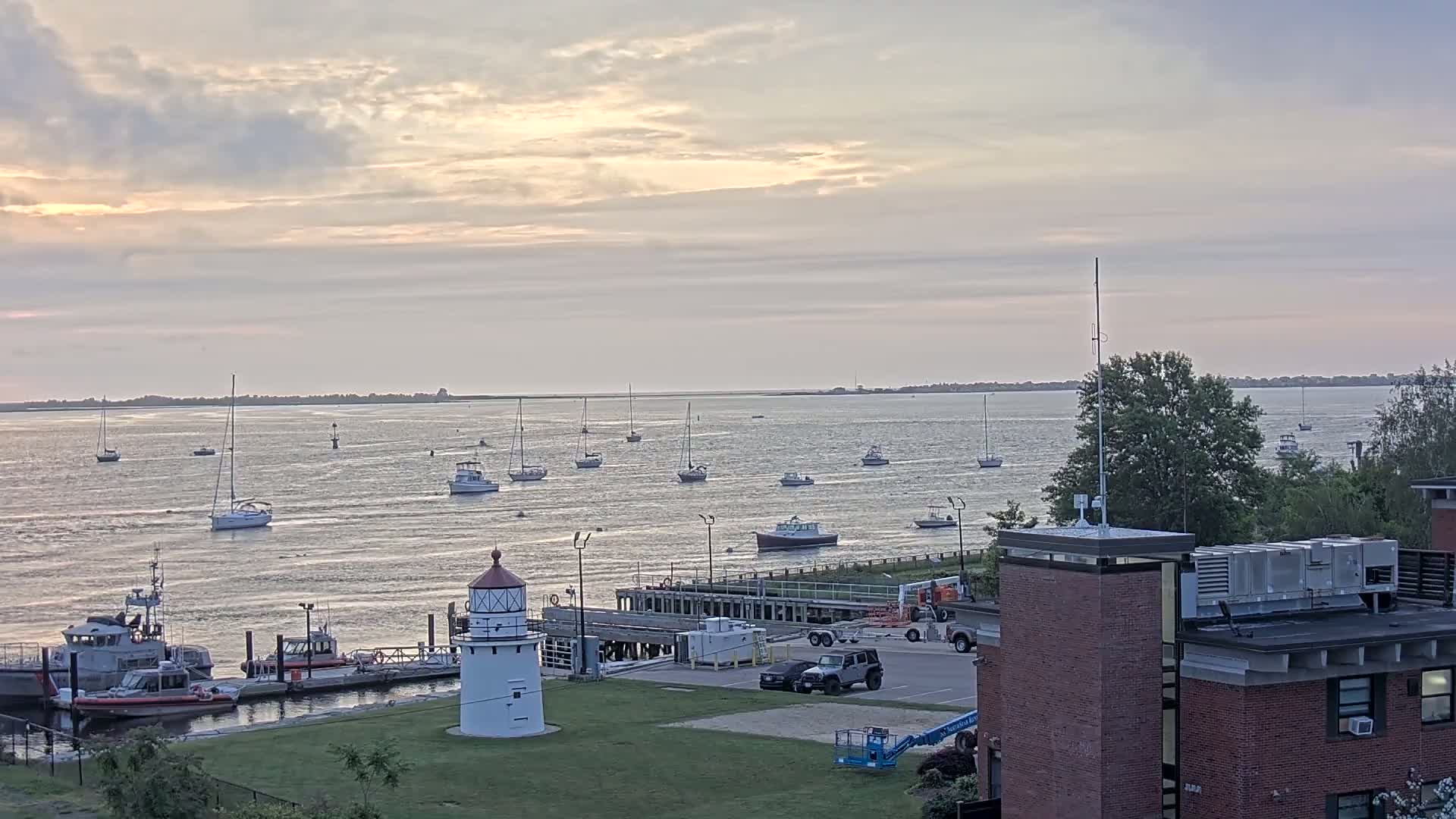A calm body of water with numerous sailboats and a few motorboats, a lighthouse, and Coast Guard vessels at a dock under a partly cloudy sunset sky.