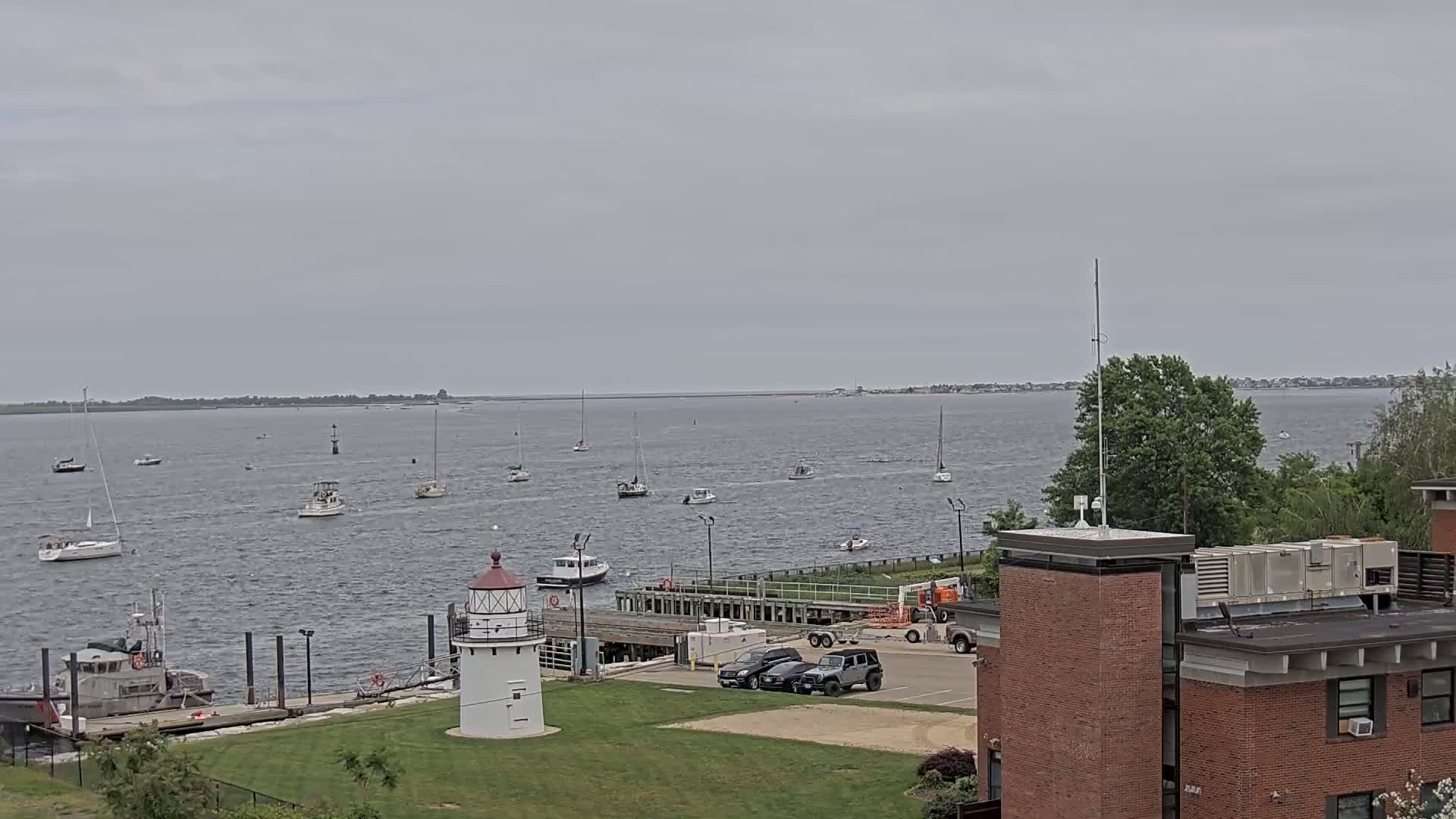 A harbor with numerous sailboats and motorboats under an overcast sky, featuring a lighthouse and a Coast Guard vessel docked near a brick building.