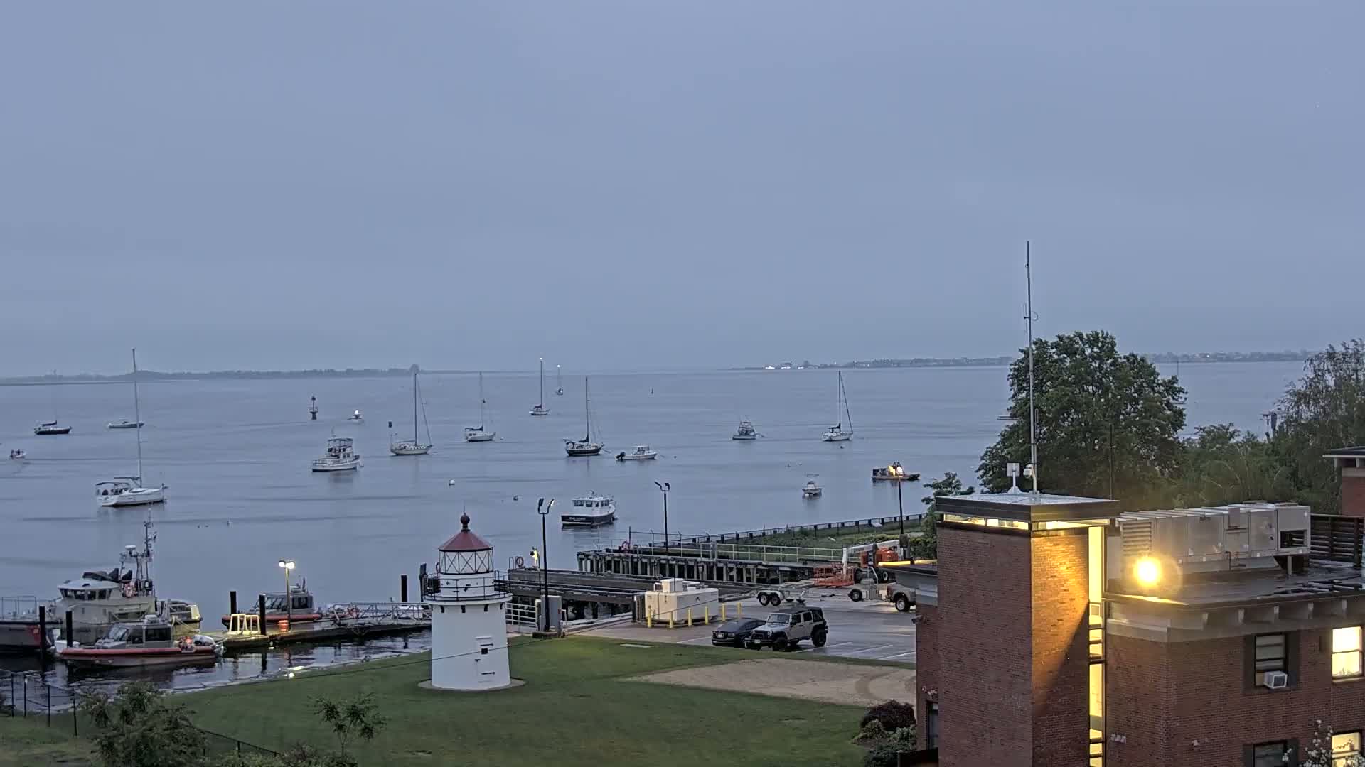 A harbor with numerous sailboats and motorboats, a lighthouse, and a brick building, under an overcast sky.