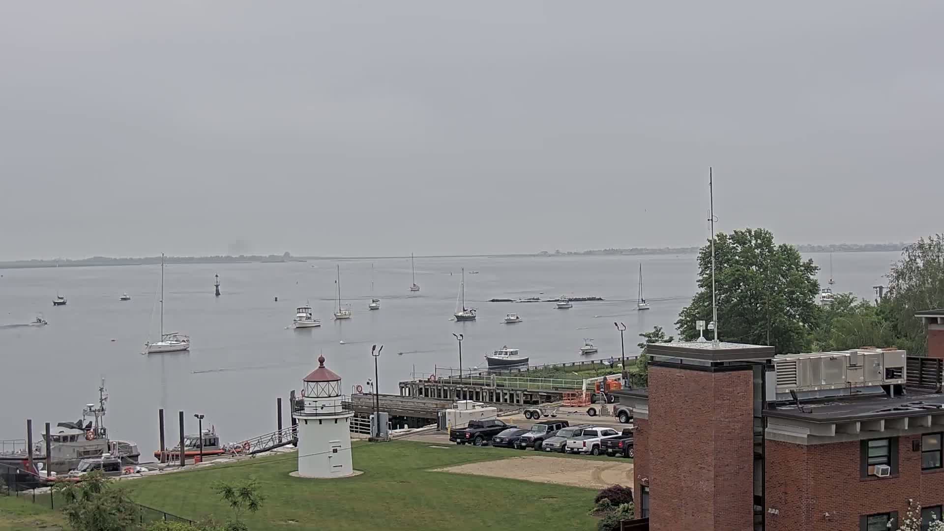 A calm, overcast day shows a harbor with several sailboats and motorboats, a lighthouse, Coast Guard vessels, and a parking lot near a brick building.
