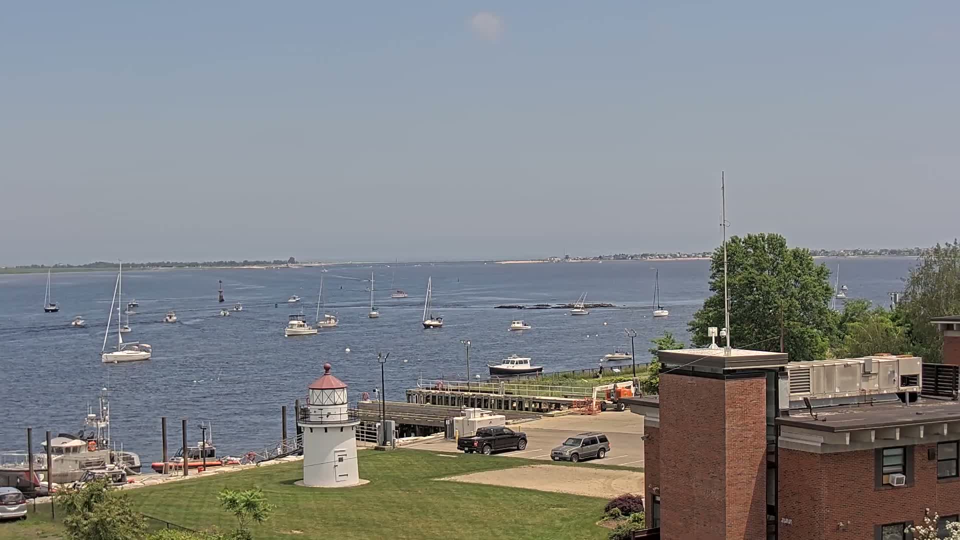 A harbor scene on a sunny day shows numerous sailboats and motorboats on calm water, a small lighthouse on a grassy area, and a brick building in the foreground.