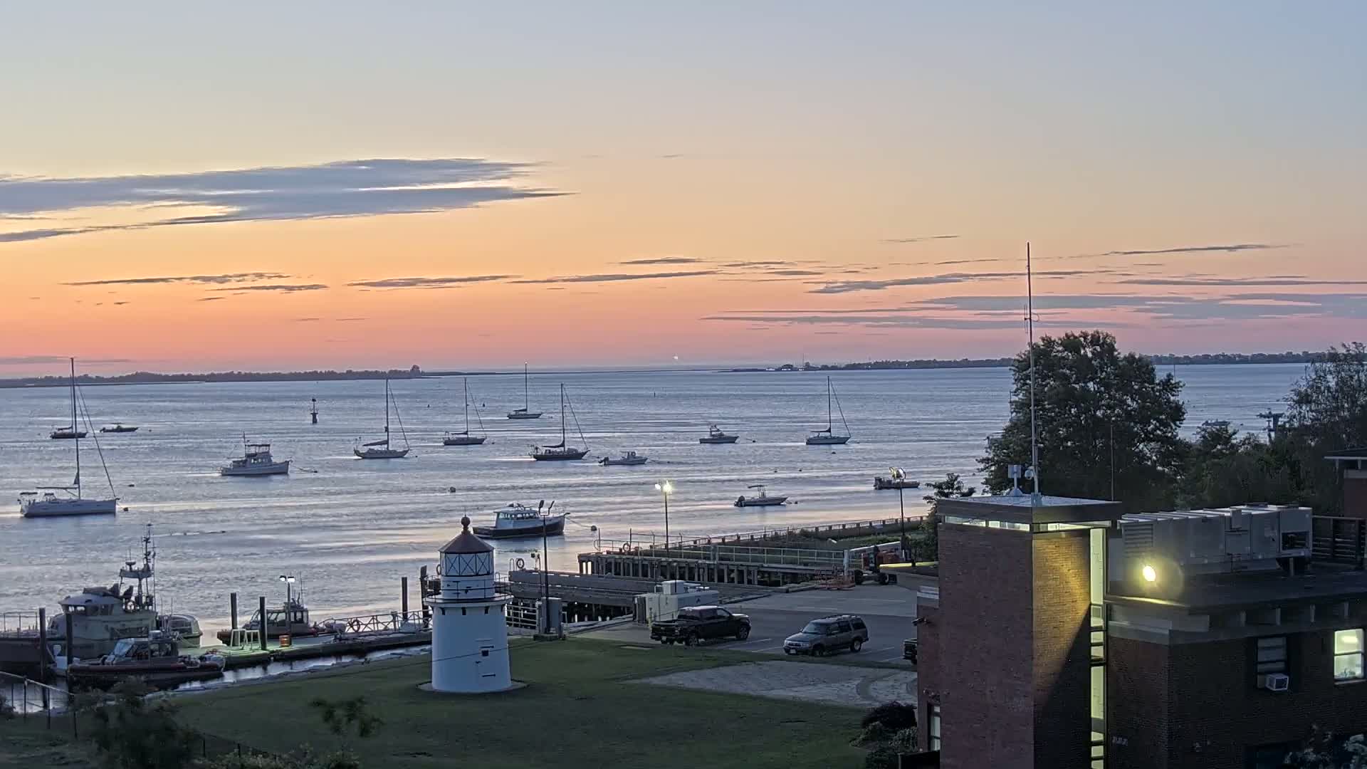 A calm body of water at sunset, dotted with sailboats and small motorboats, is observed from a vantage point overlooking a harbor with a lighthouse, a dock, and a brick building.