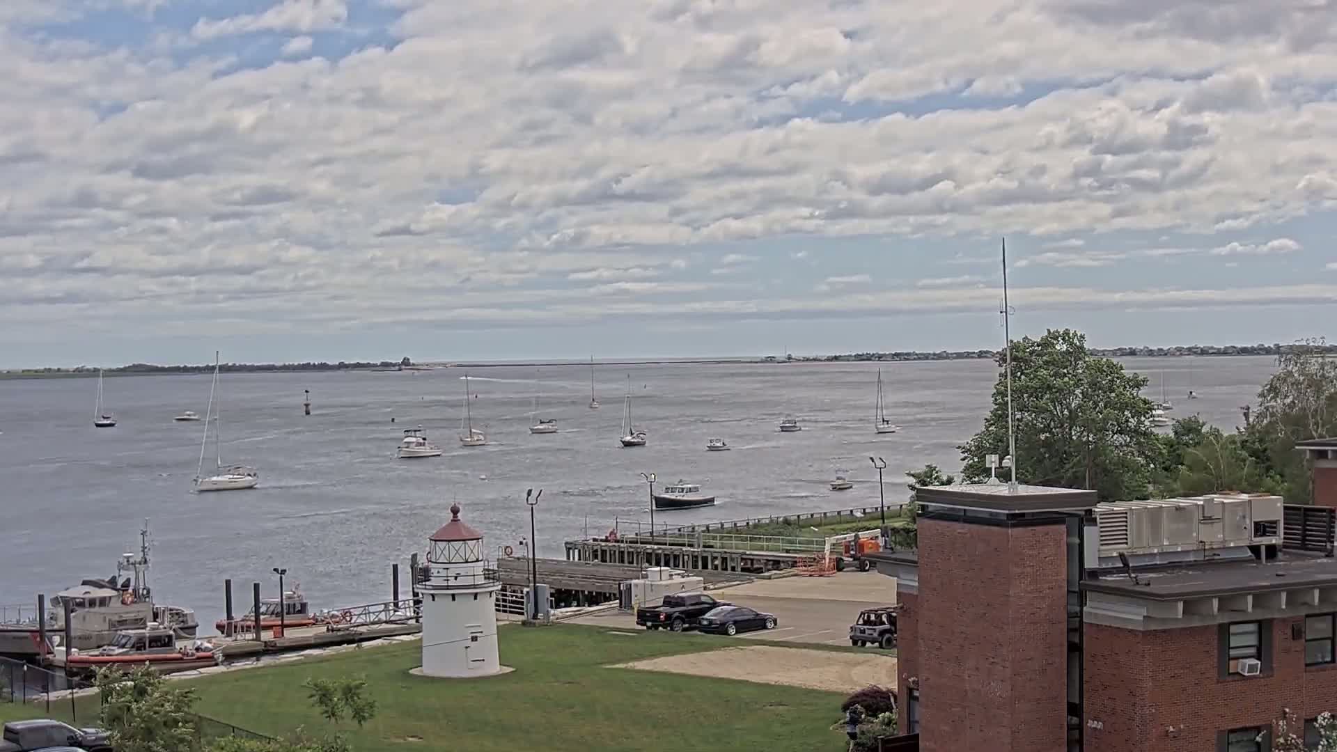 A partly cloudy day shows a calm body of water with numerous sailboats and motorboats, a lighthouse, and a Coast Guard station; a brick building is in the foreground to the right.