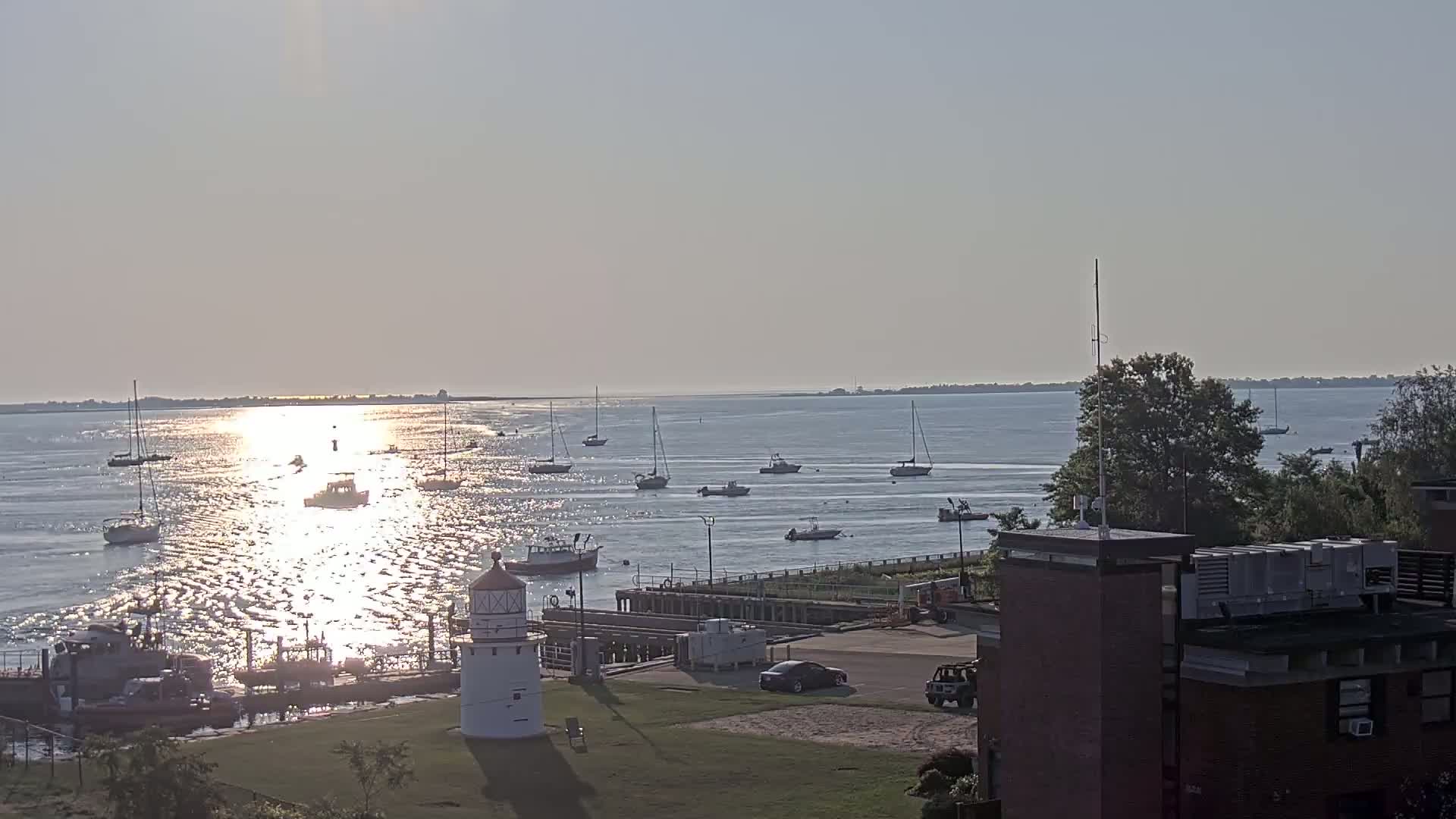 A calm, sunny day shows a harbor with numerous boats, a lighthouse, and a building in the foreground.