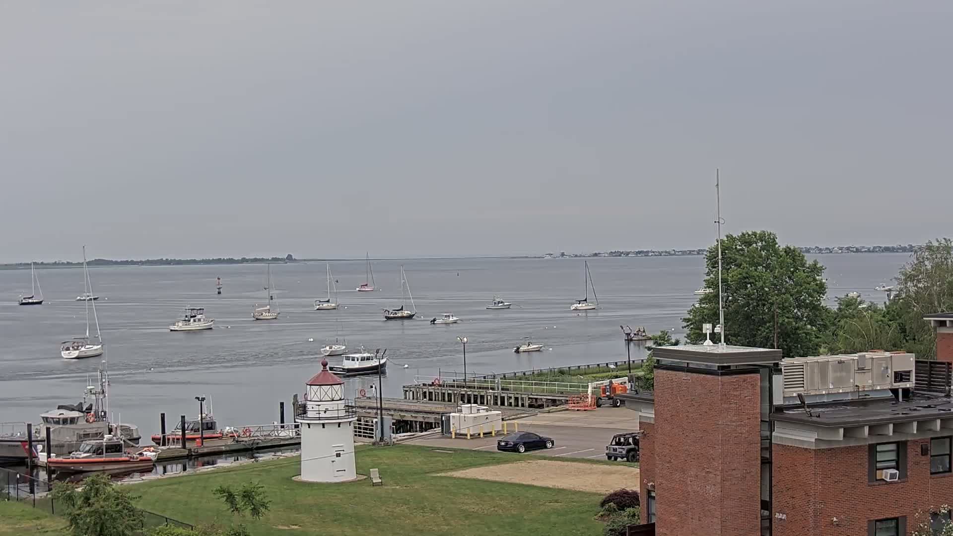 A calm, overcast day shows a harbor with numerous sailboats and motorboats, a lighthouse, Coast Guard vessels docked, and a brick building in the foreground.