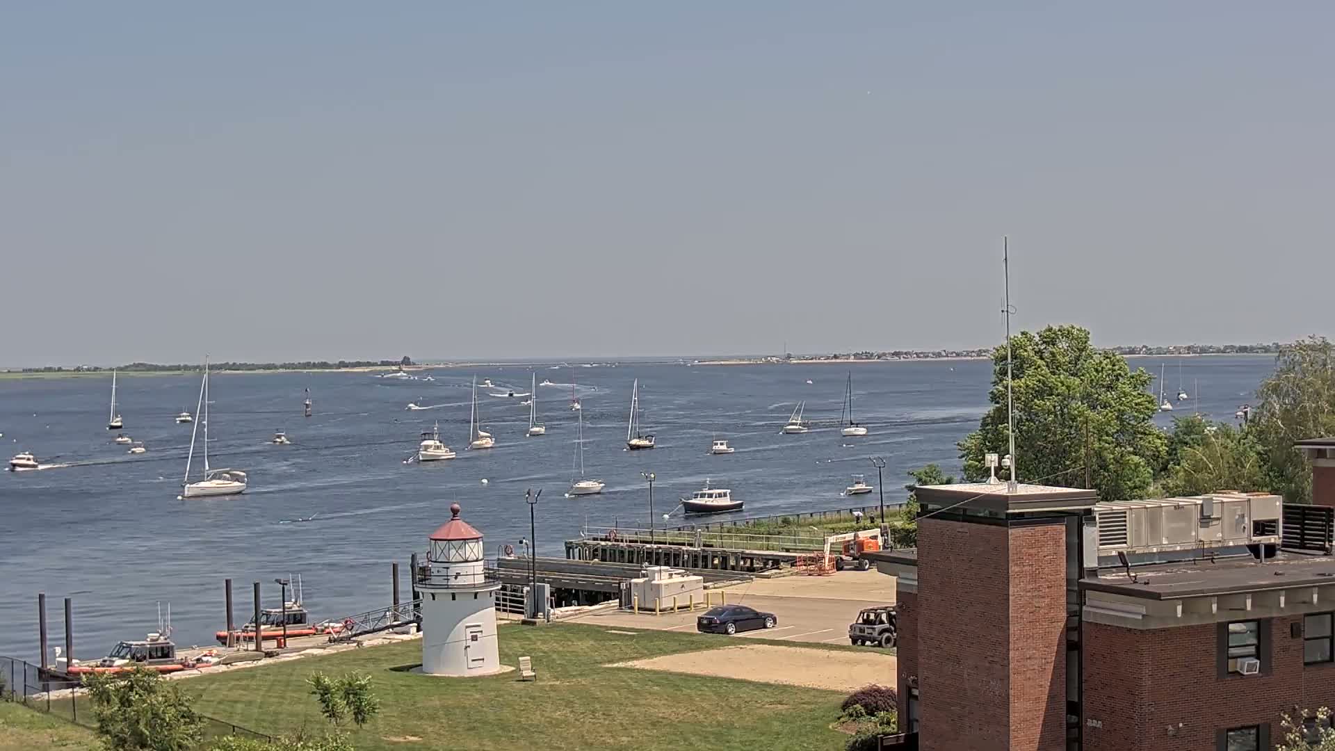 A white lighthouse stands on a grassy area overlooking a calm bay with numerous sailboats and motorboats on a sunny day.
