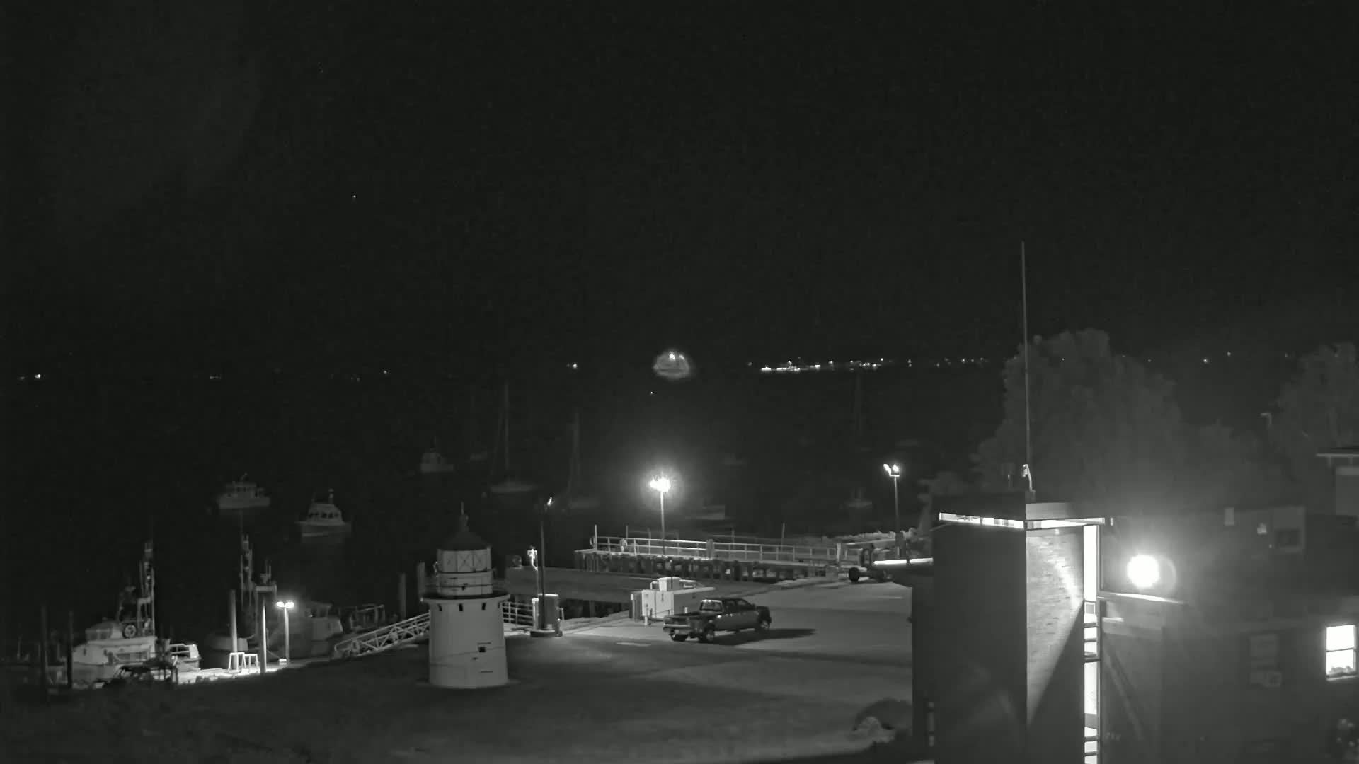 A nighttime view of a harbor with several boats docked, a small lighthouse, a pickup truck, and a building under a dark, clear sky.