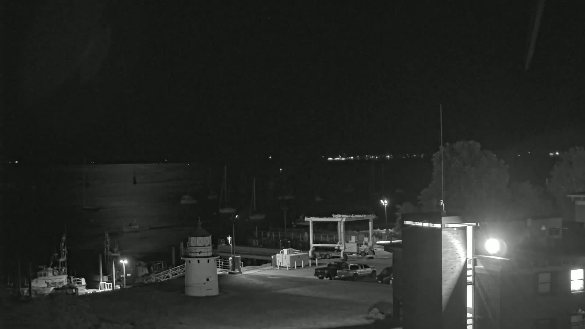 A nighttime view of a harbor with boats, a lighthouse, and several vehicles parked near a building under a clear, dark sky.