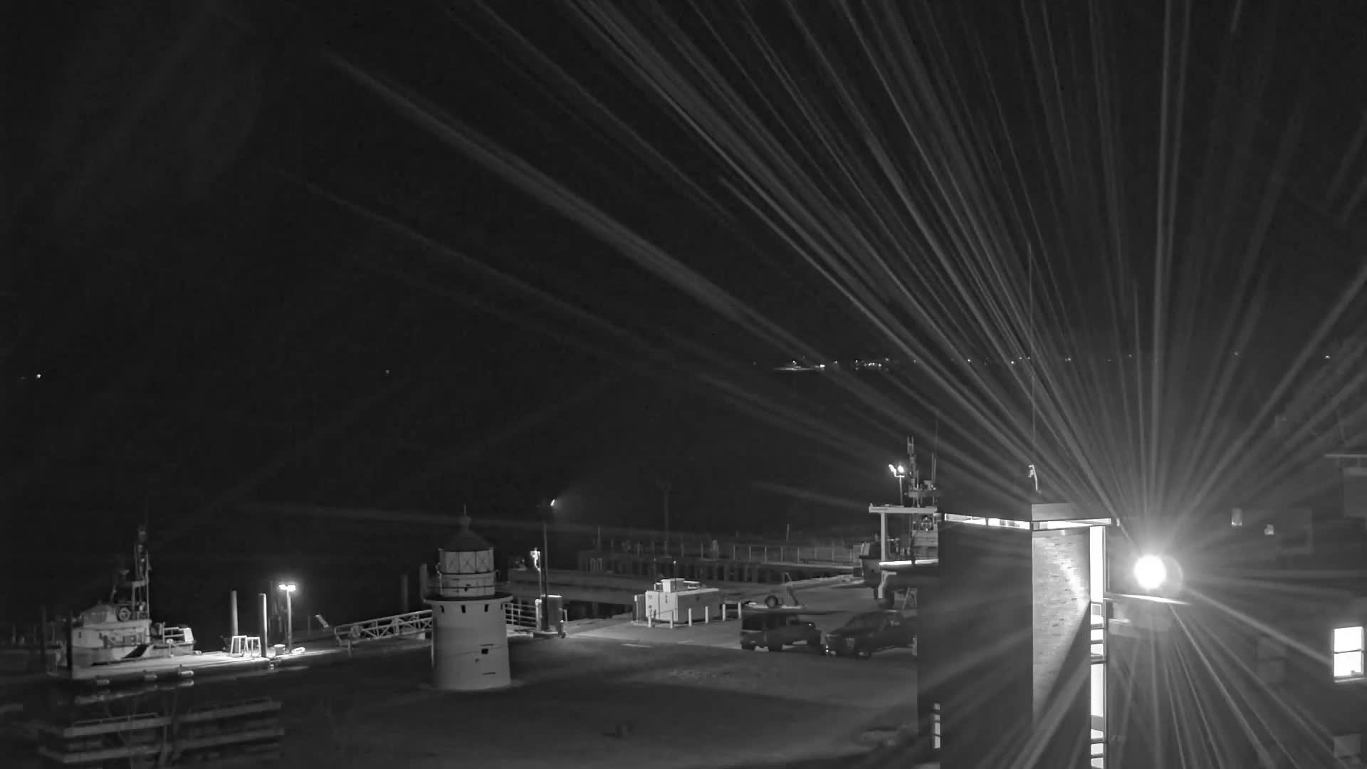 A misty or foggy nighttime scene captures a harbor featuring a lighthouse, boats docked at a pier, and several vehicles, all under the glow of streetlights and a powerful, radially streaking light source on the right.