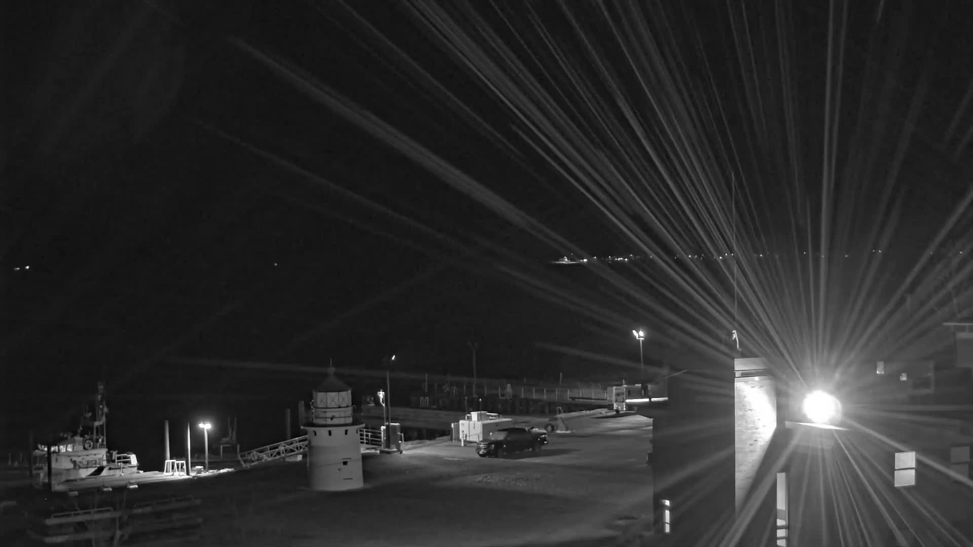 A clear night captured in black and white reveals a harbor with a lighthouse, a docked vessel, and a parked car on a pier, brightly illuminated by lights causing significant lens flares that stretch across the dark sky.