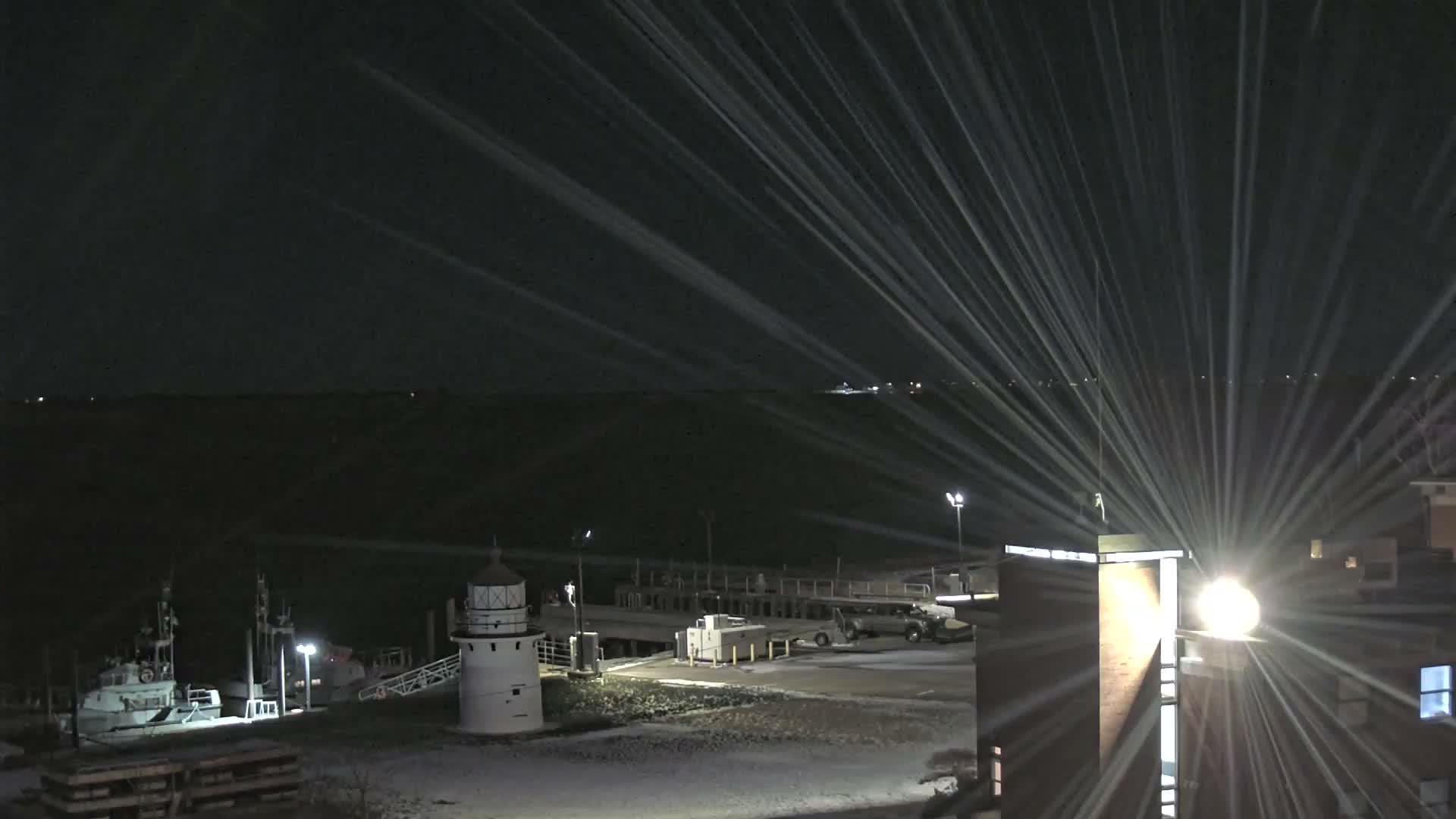 A dark, snowy night scene depicts a white lighthouse and a docked boat by a pier, with bright artificial lights creating prominent streaks as snow falls and covers the ground.