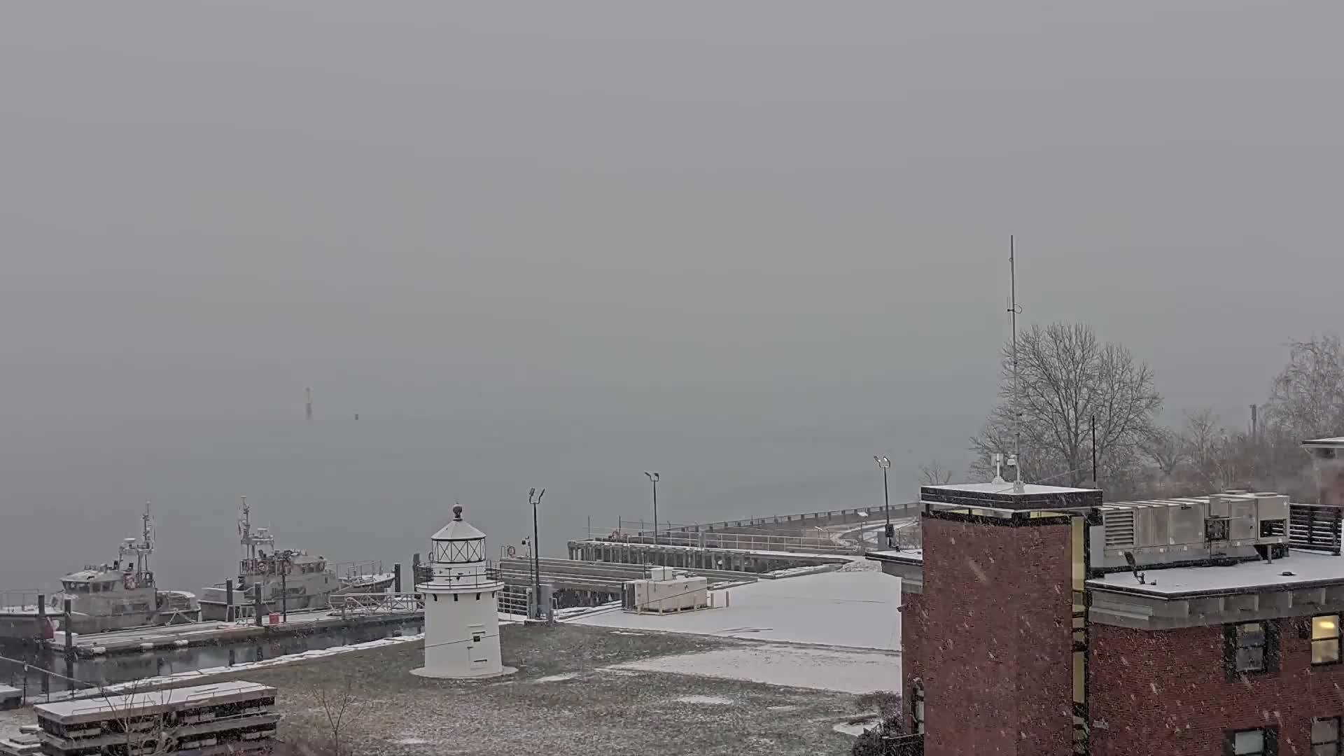 A dark, snowy night scene depicts a white lighthouse and a docked boat by a pier, with bright artificial lights creating prominent streaks as snow falls and covers the ground.